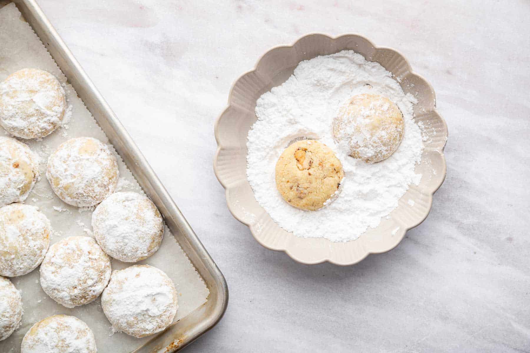 Two cookies being rolled in powdered sugar in a small dish.