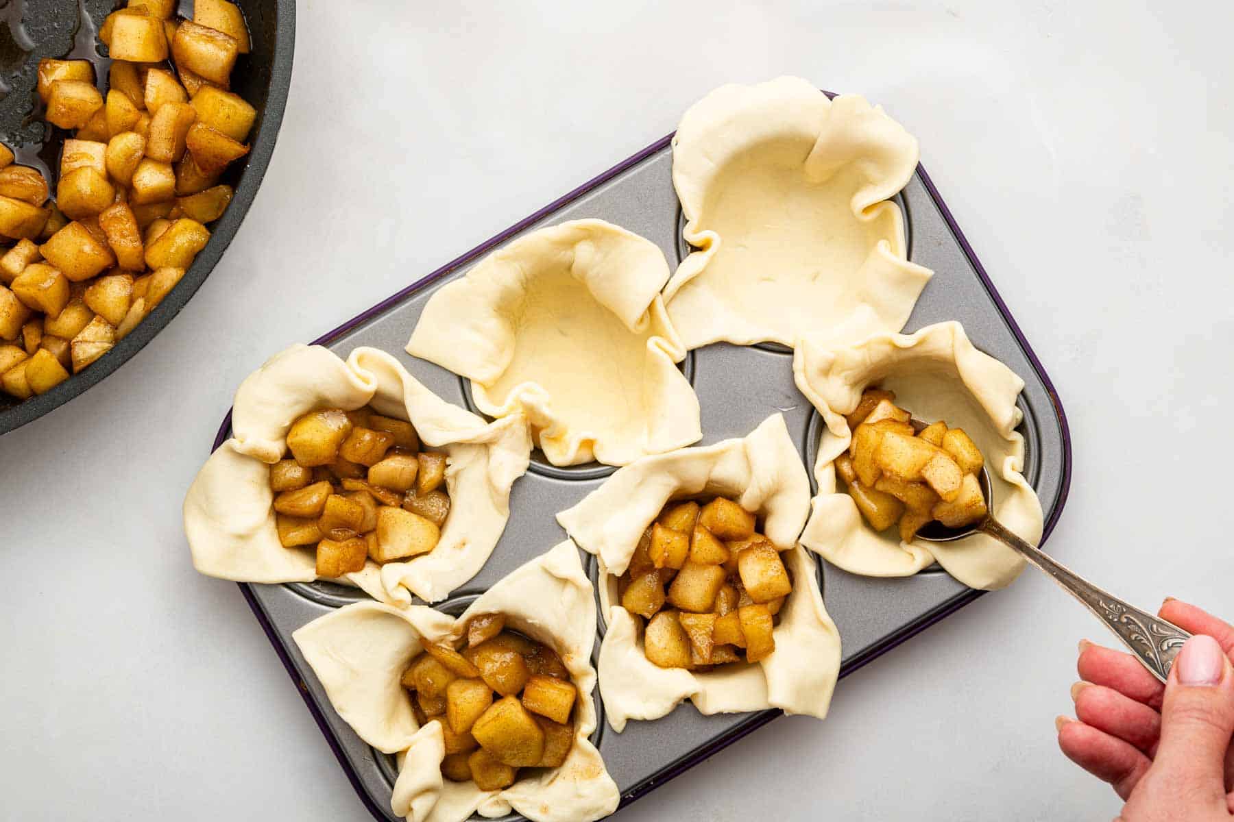 Hand spooning apple pie filling into puff pastry cups in a muffin pan.