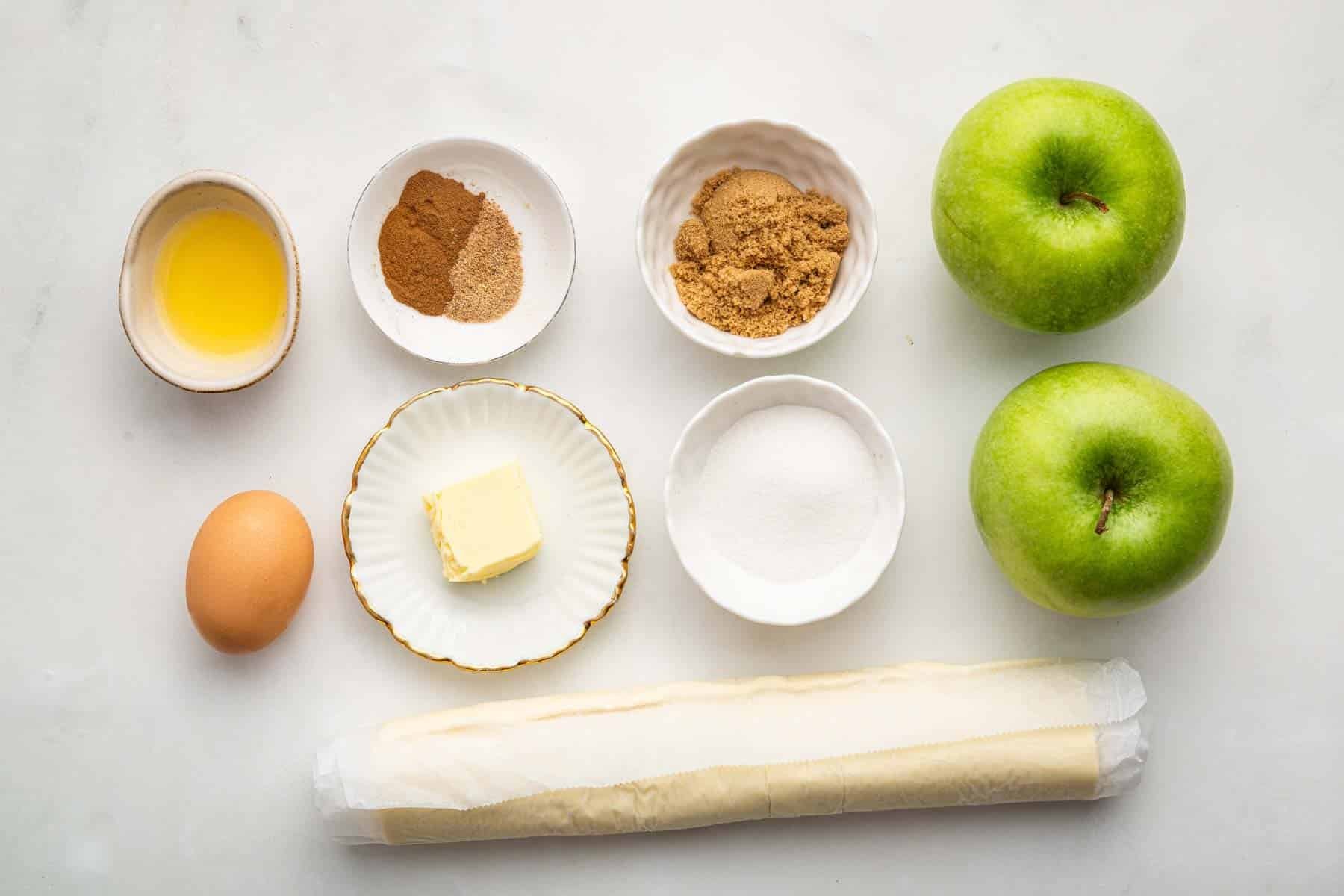 Puff pastry, two green apples, flour, butter and spices in bowls on counter.