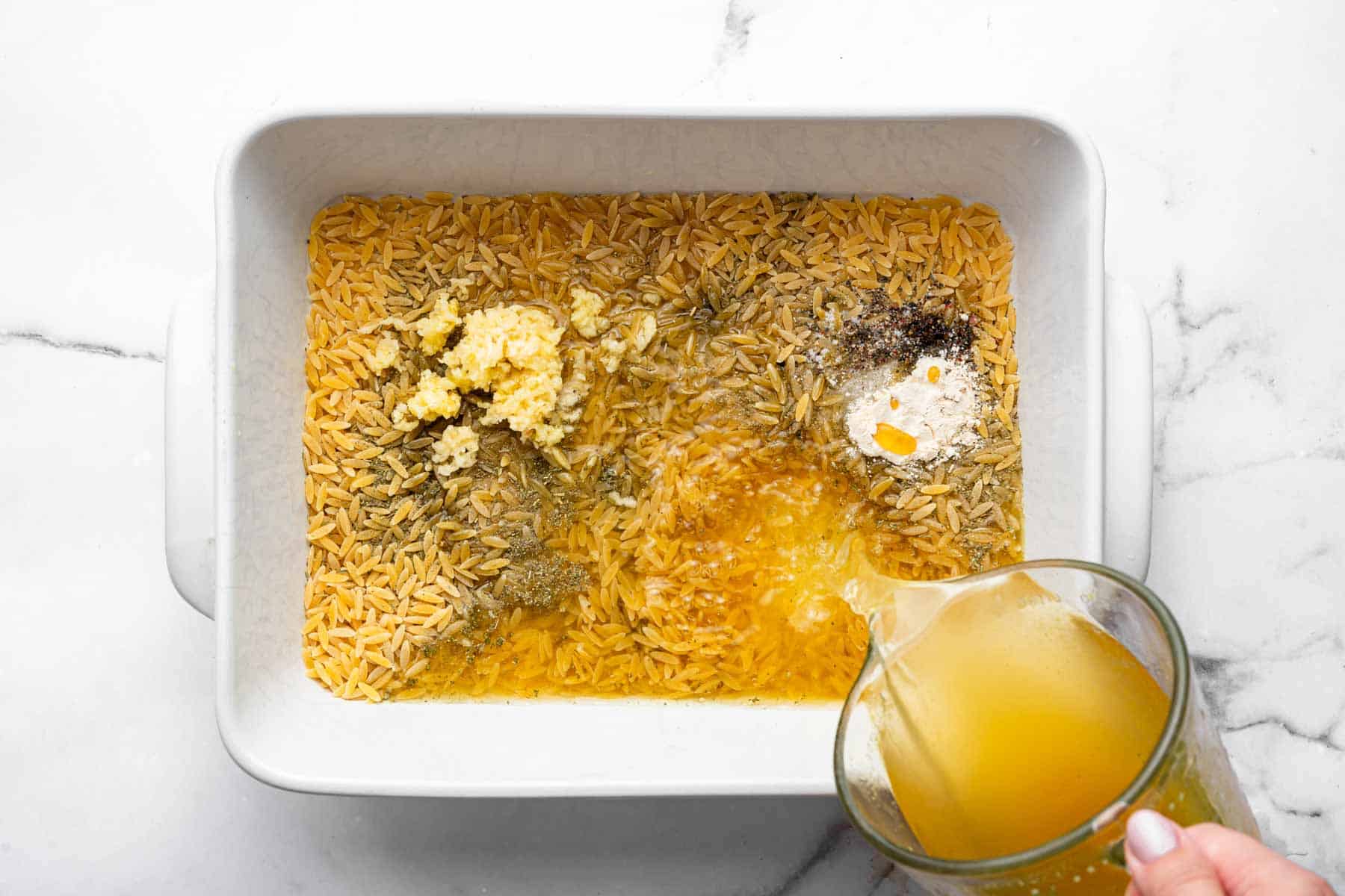 Pouring broth in a rectangular baking dish with pasta and herbs.