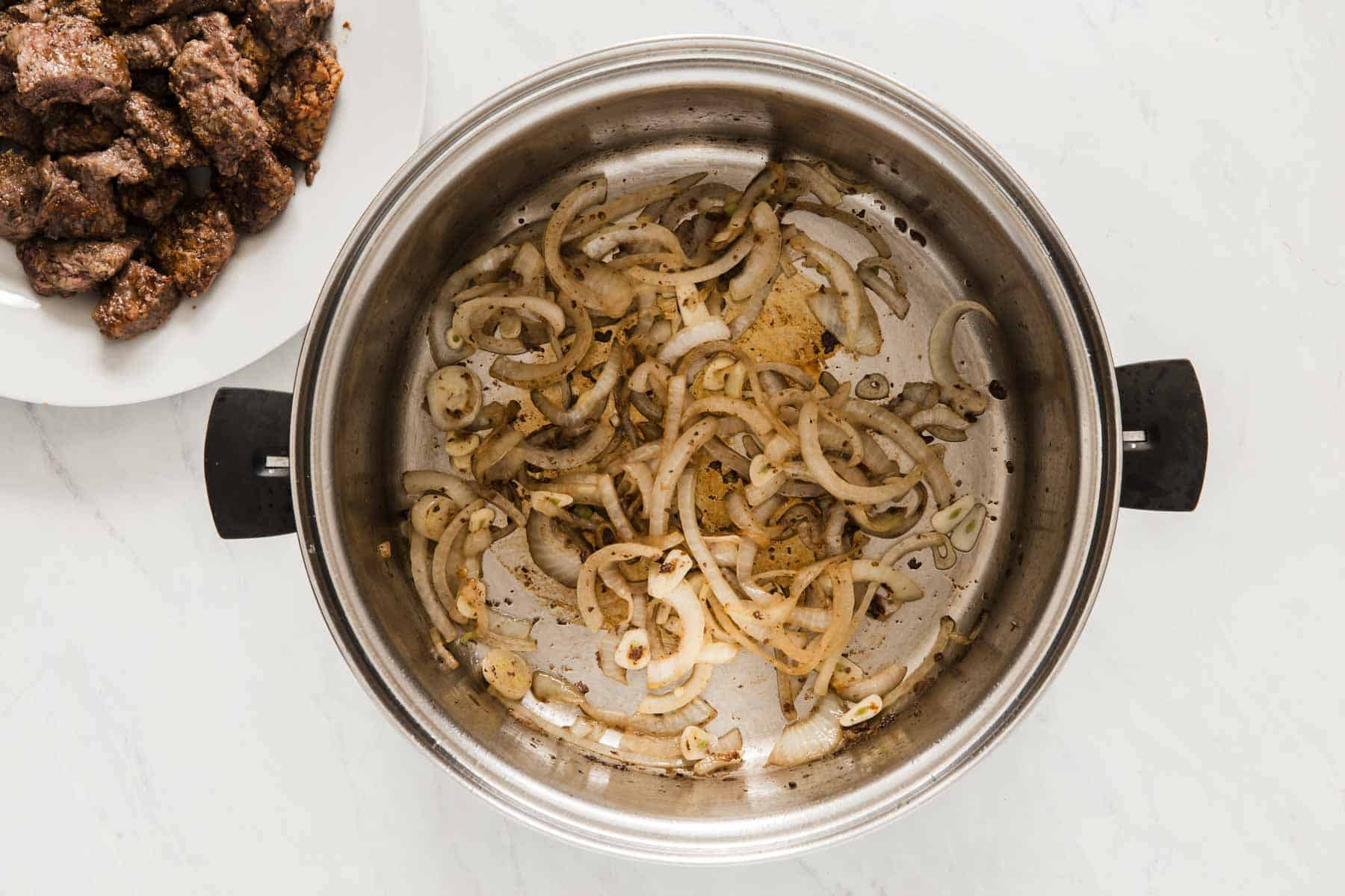 Sliced onions cooking in a pan with beef stew meat on the side on a plate.