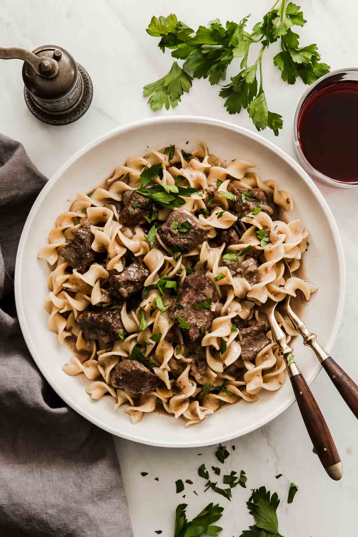 Overhead lay-flat shot of noodles and beef in white bowl with serving spoons.