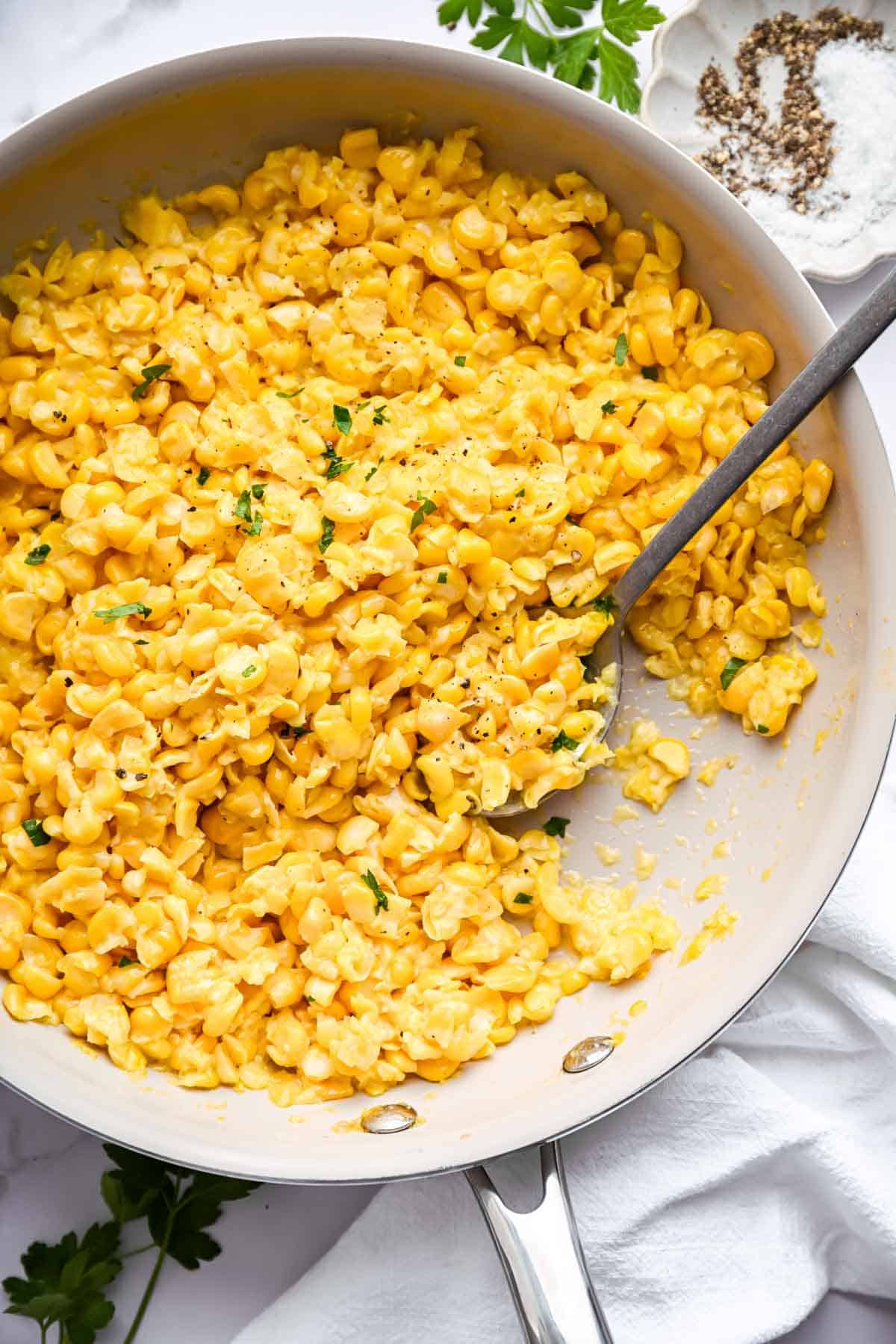 White skillet of buttered corn on table with parsley garnish.