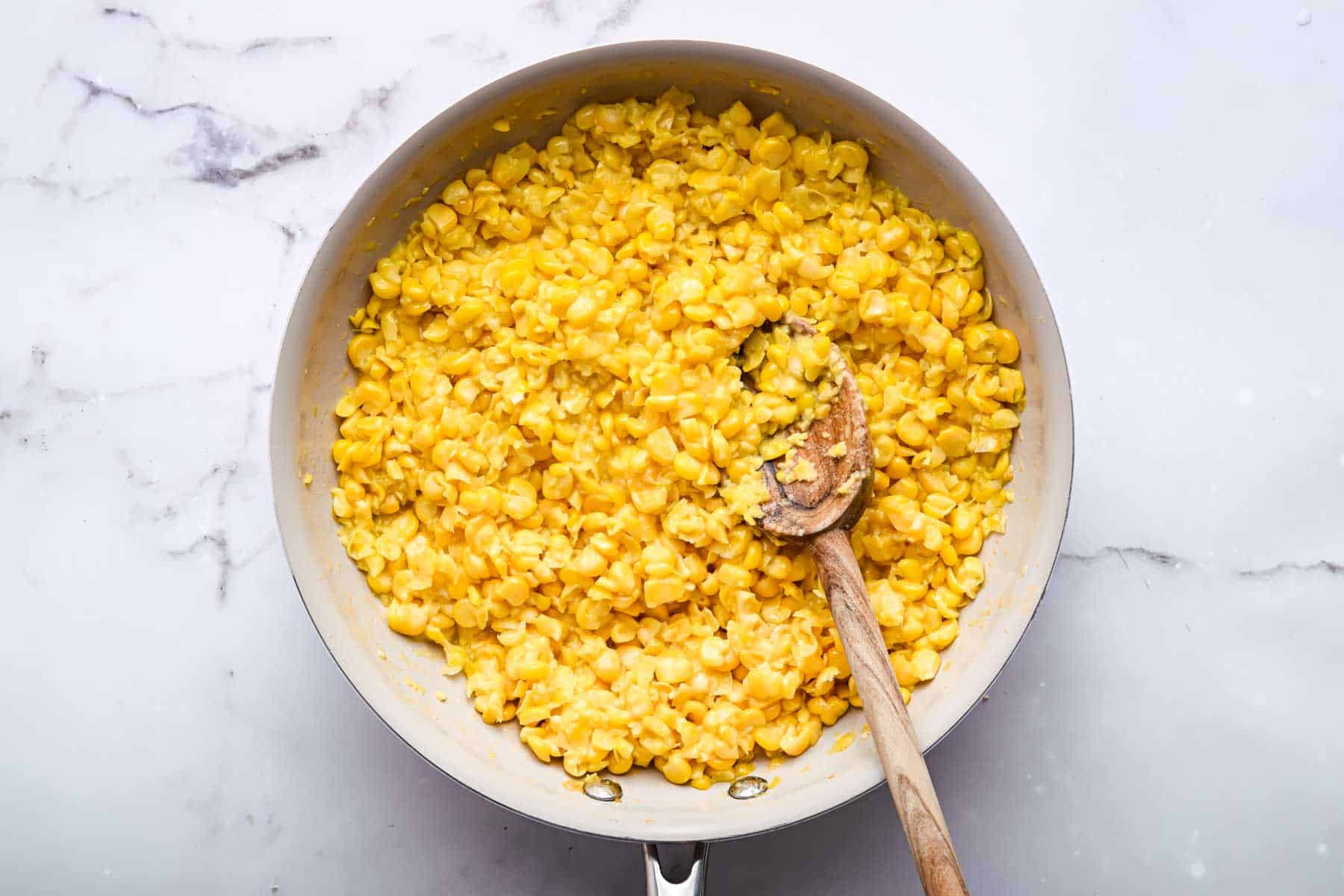 Bright yellow vegetables in white skillet with wooden spoon.