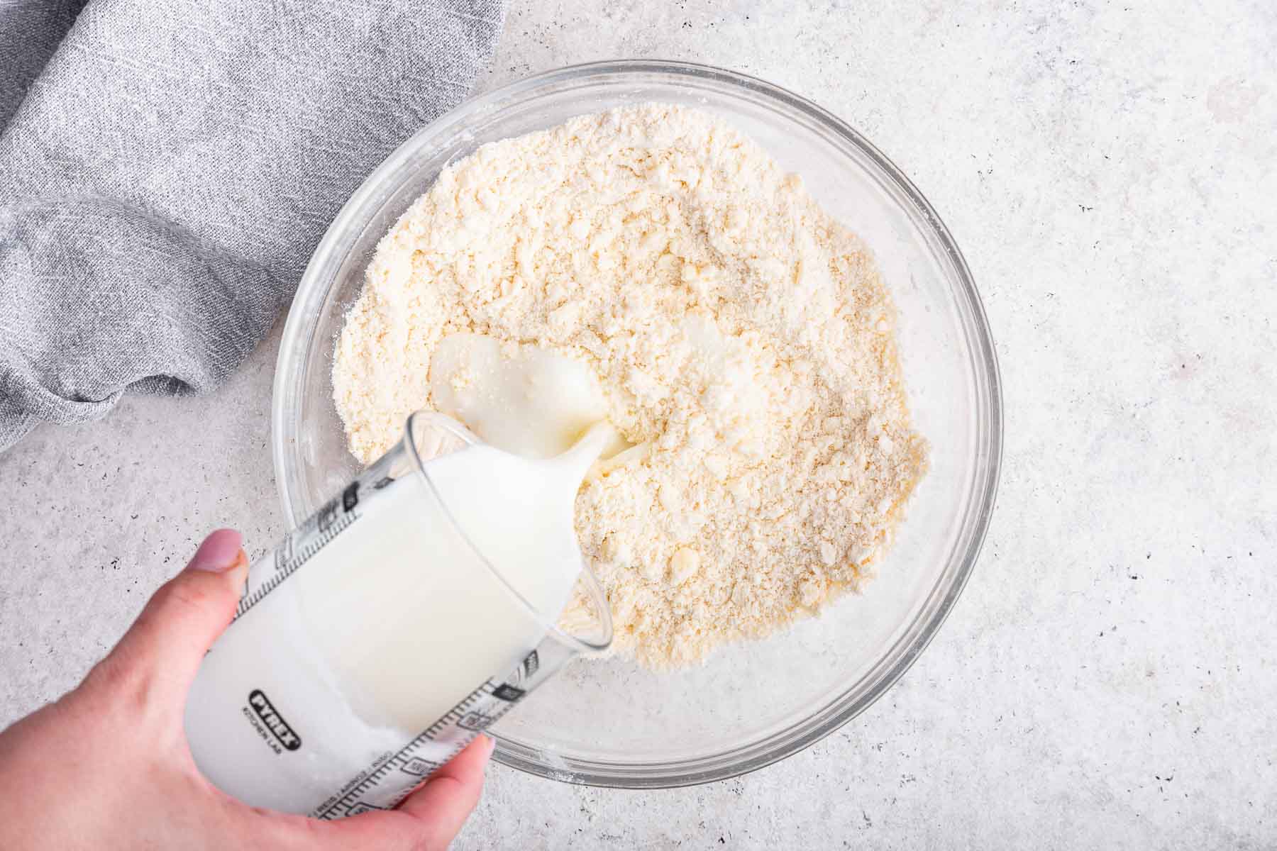 Hand pouring buttermilk into bowl with flour and butter.