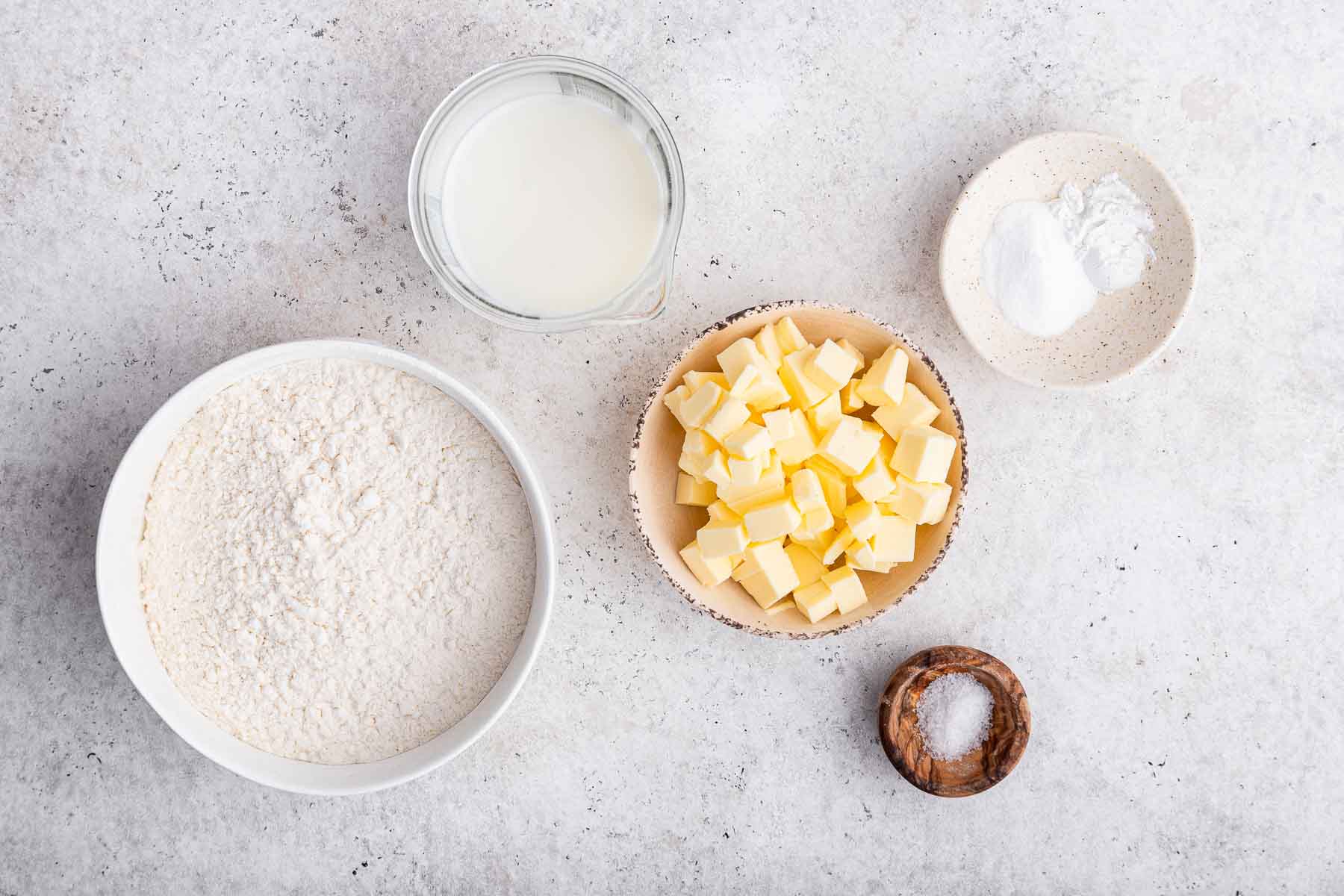 Overhead shot of flour, cubed butter, salt and baking soda on grey counter.
