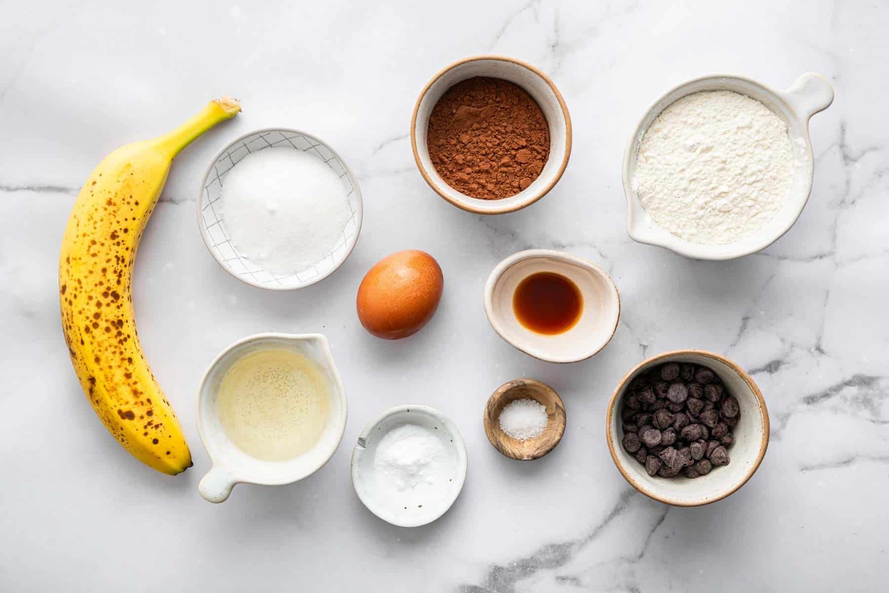 Small bowls of flour, sugar, chips, cocoa powder and fruit on a marble counter.