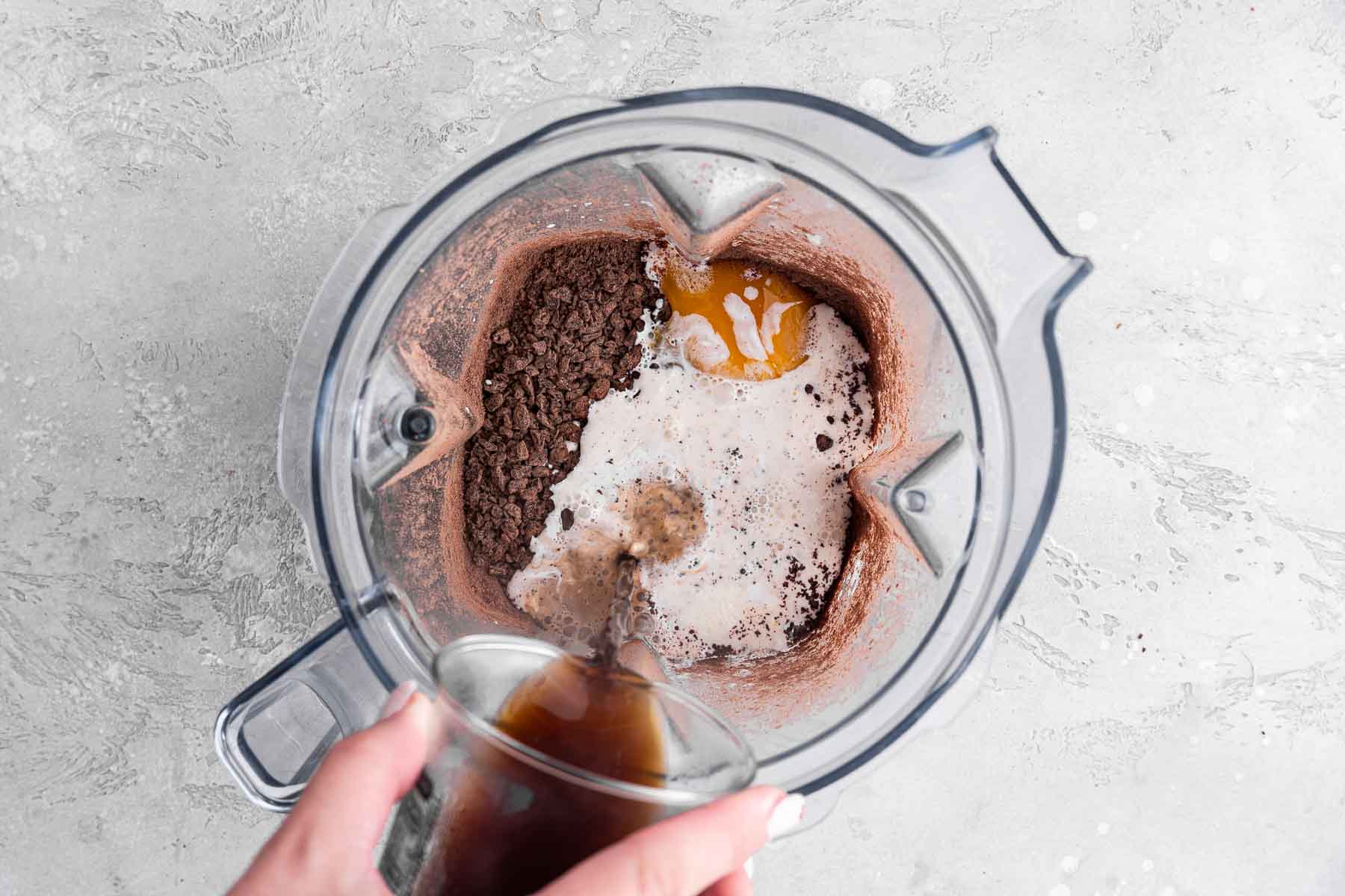 Pouring hot espresso in a blender with white liquid and brown pieces.