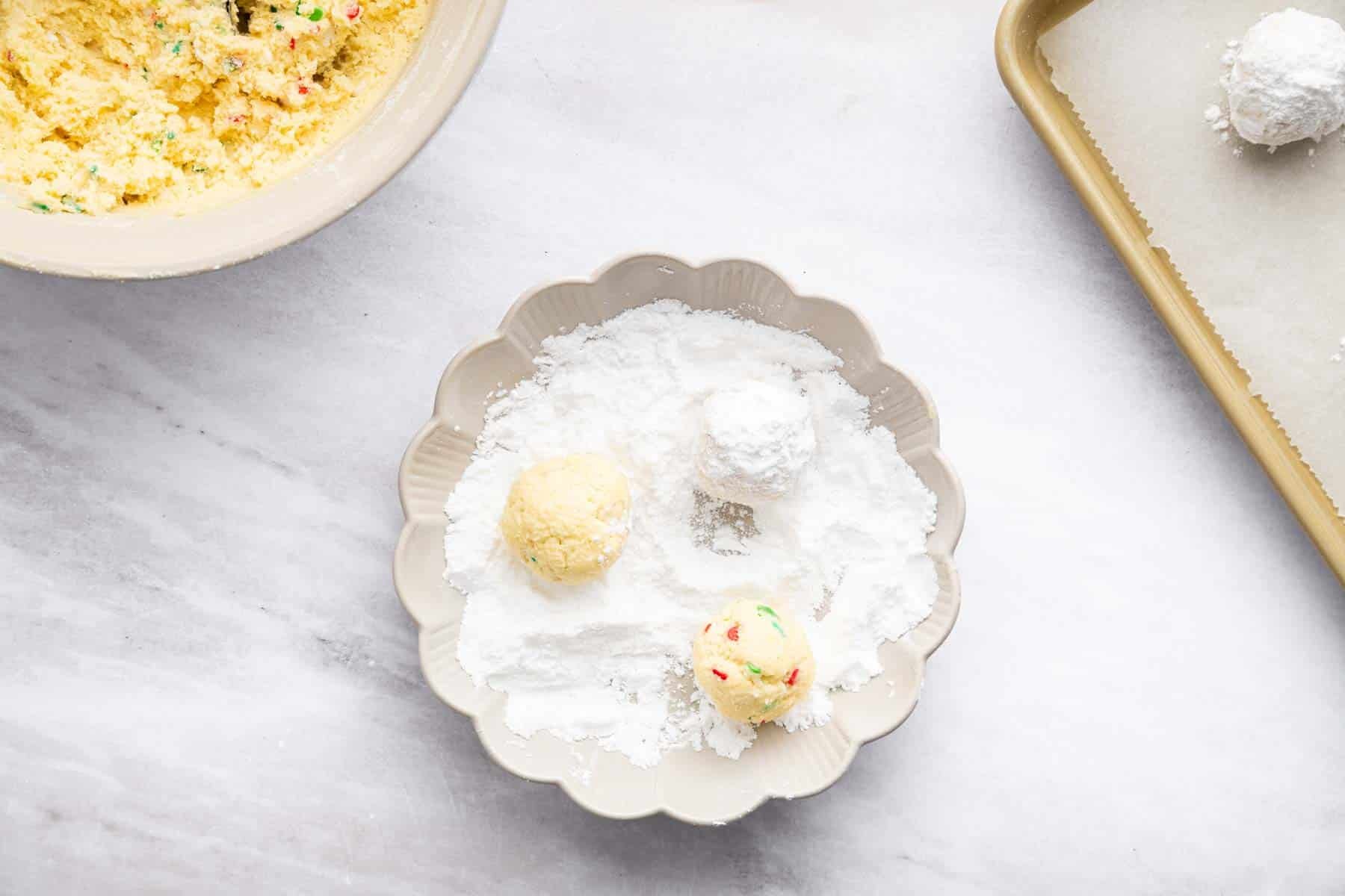 Rolling three yellow cookie dough balls in powdered sugar in shallow bowl.