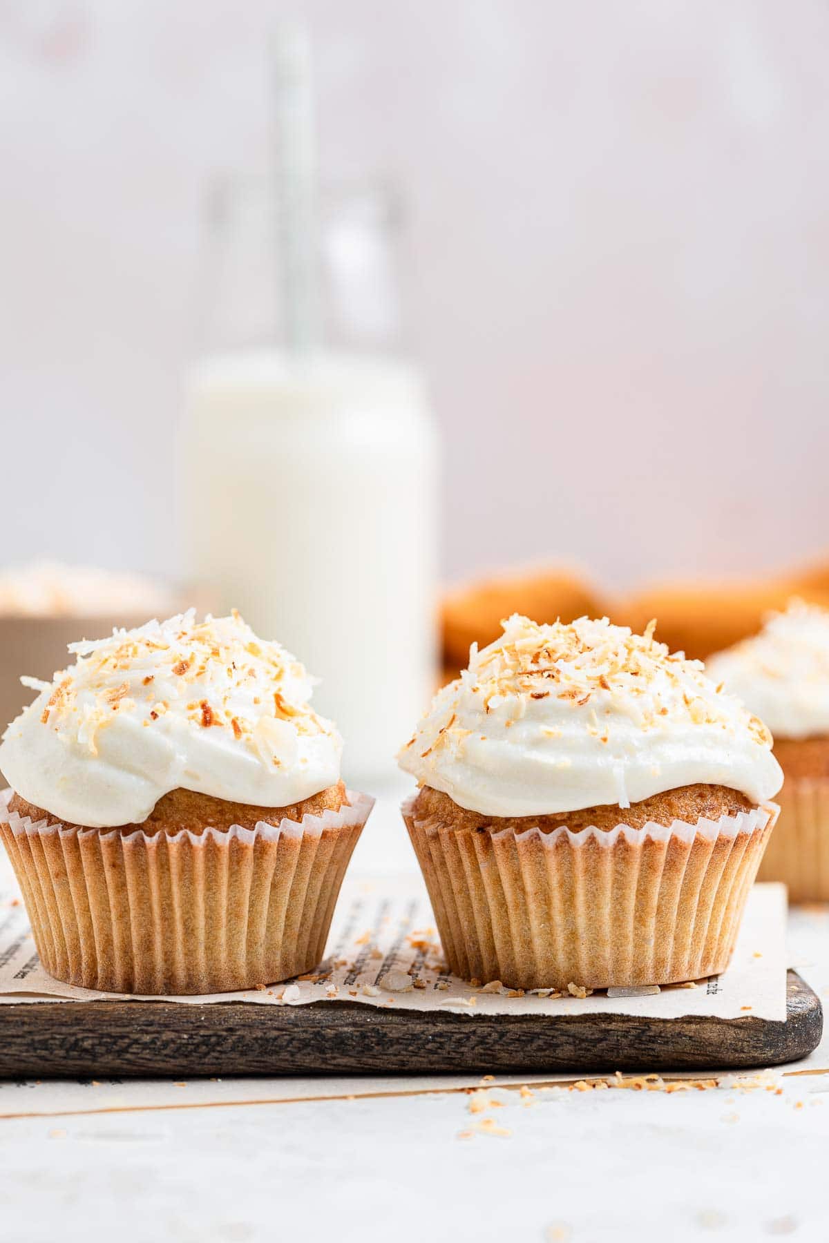 Two frosted coconut cupcakes with milk in the background.