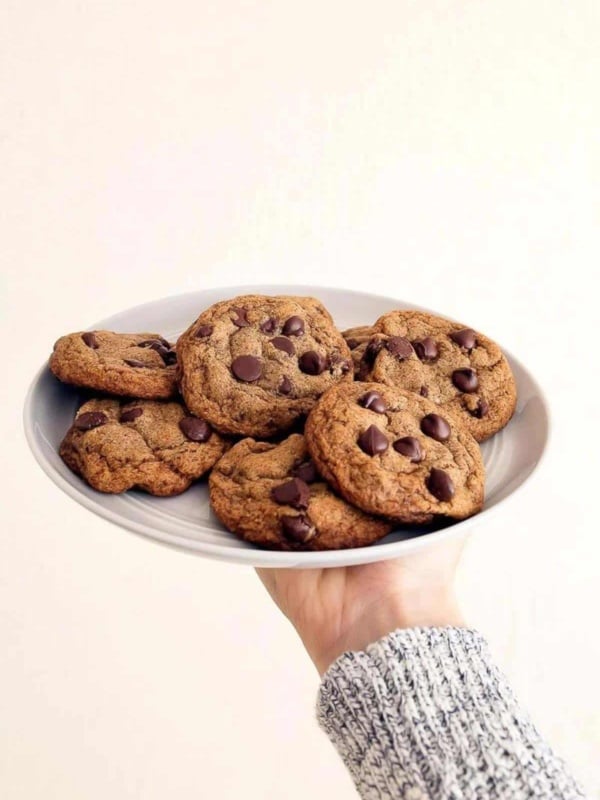 Arm wearing sweater holding up a grey plate of healthy chocolate chip cookies.