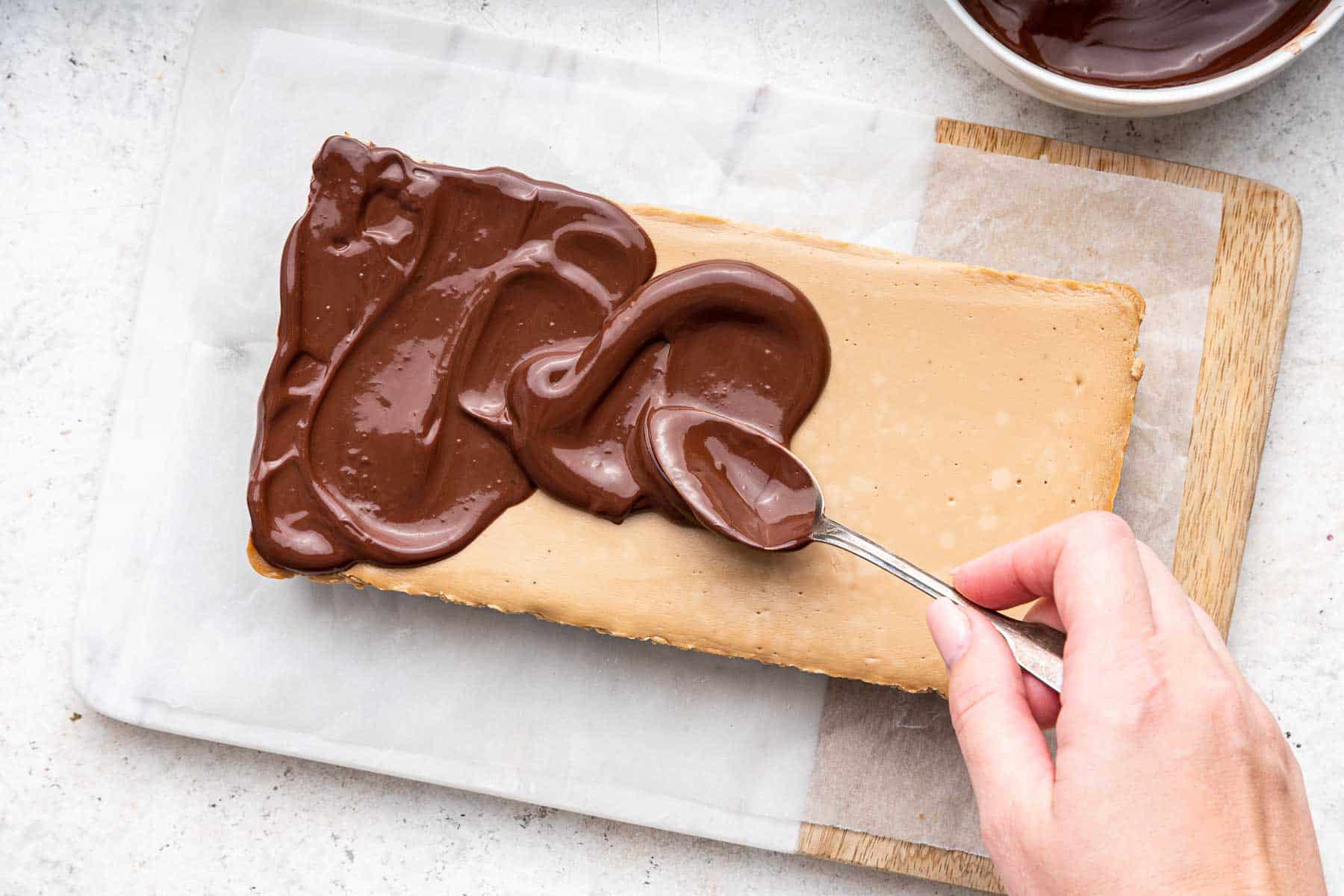 Hand spreading chocolate ganache on brown rectangular dessert.