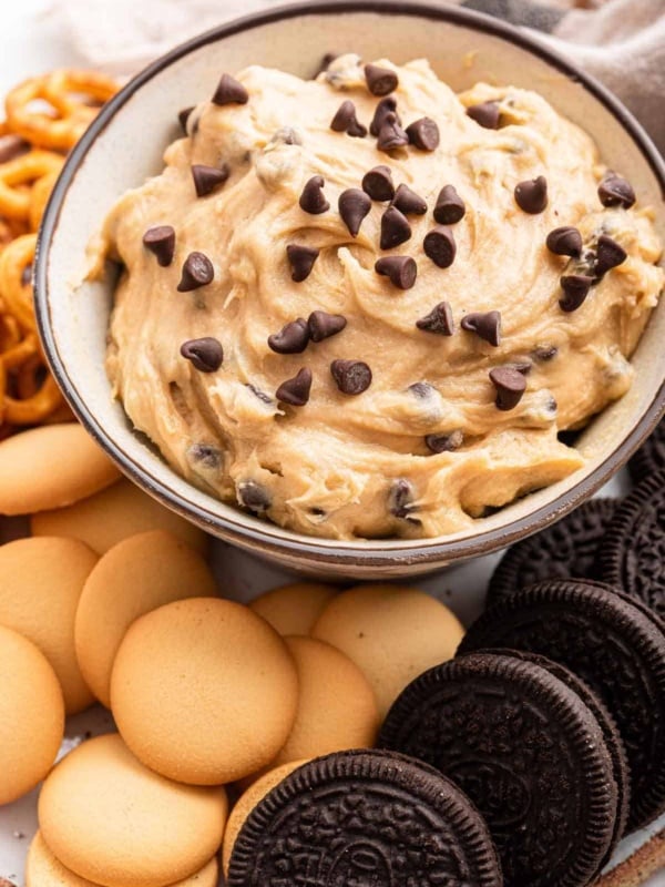 Vertical image of cookie dough dip in white bowl surrounded by cookies.