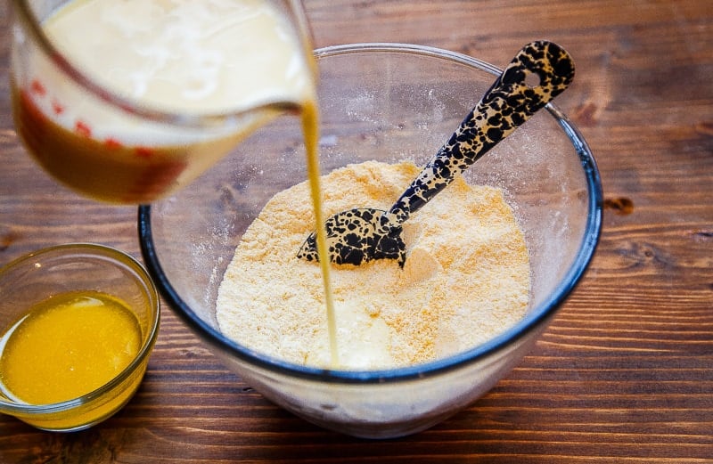 Pouring milk into bowl of yellow batter with spoon inside.