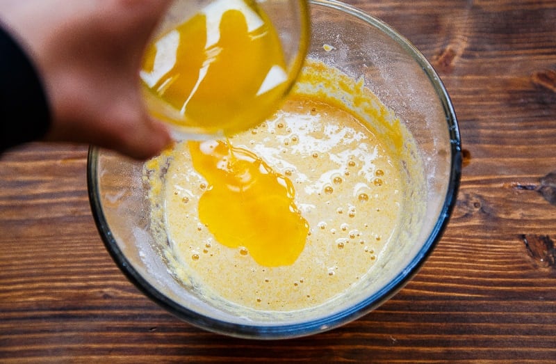 Hand pouring melted butter into glass bowl with yellow batter.