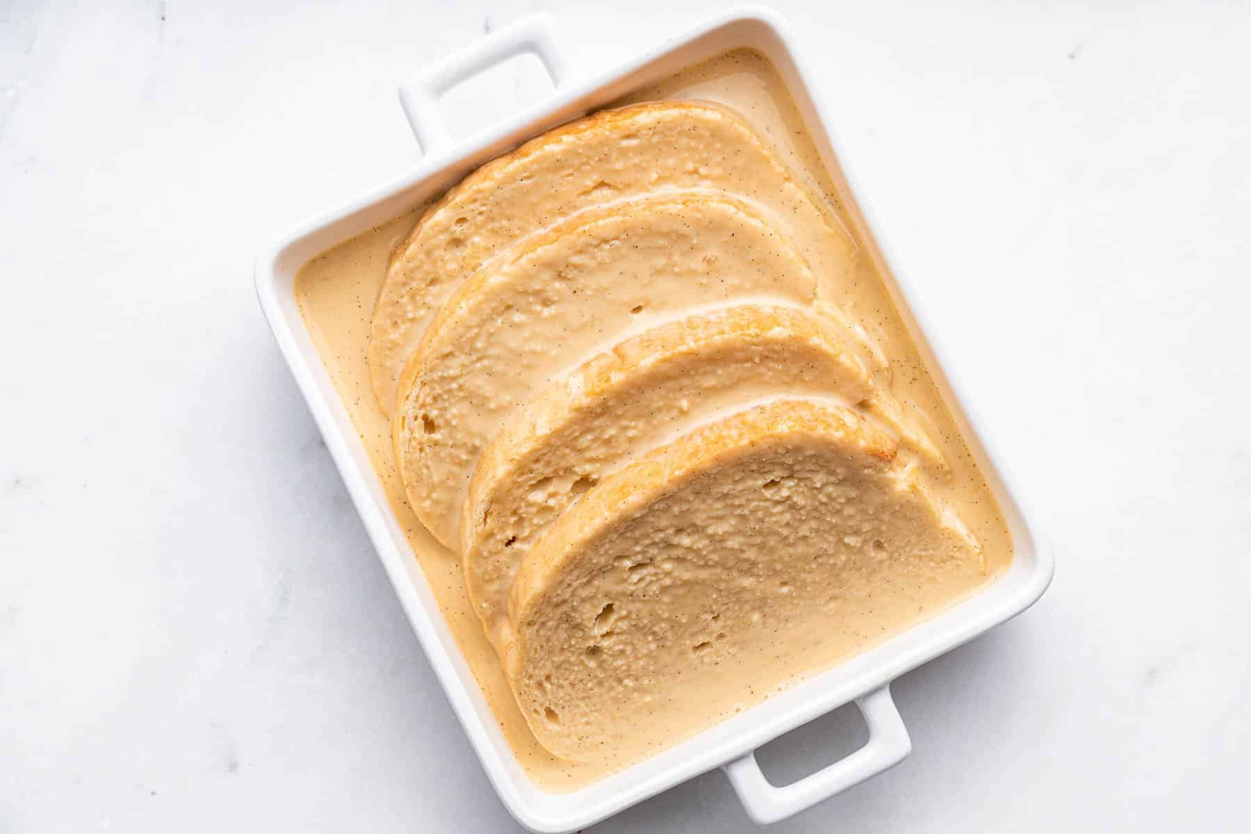 Four big slices of bread in a white baking dish with custard poured on top.