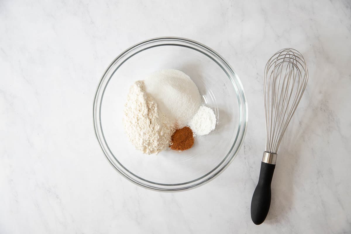 Clear bowl with flour, baking powder, nutmeg, and salt and a whisk on the side.