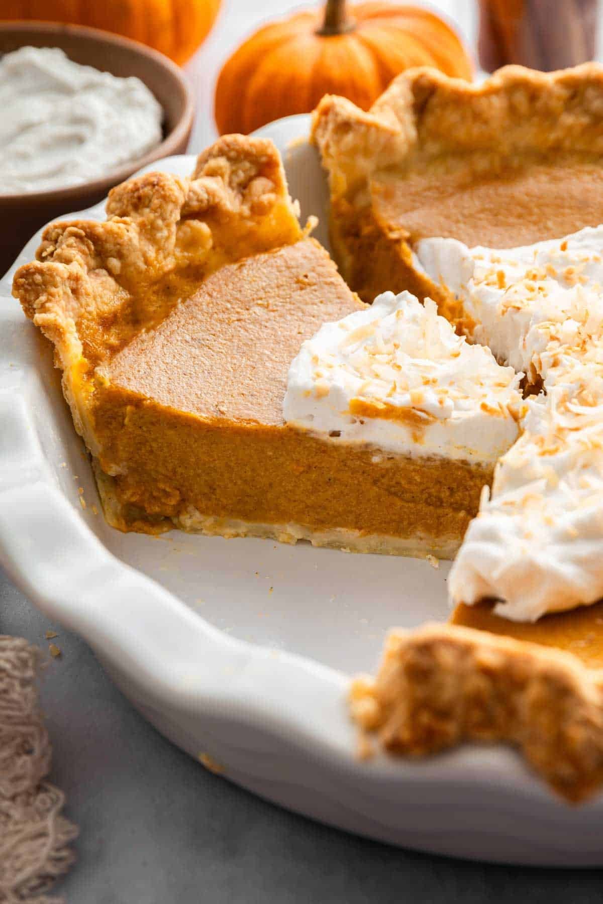 Slice of dairy free pumpkin pie in pie plate with coconut whipped cream on top.