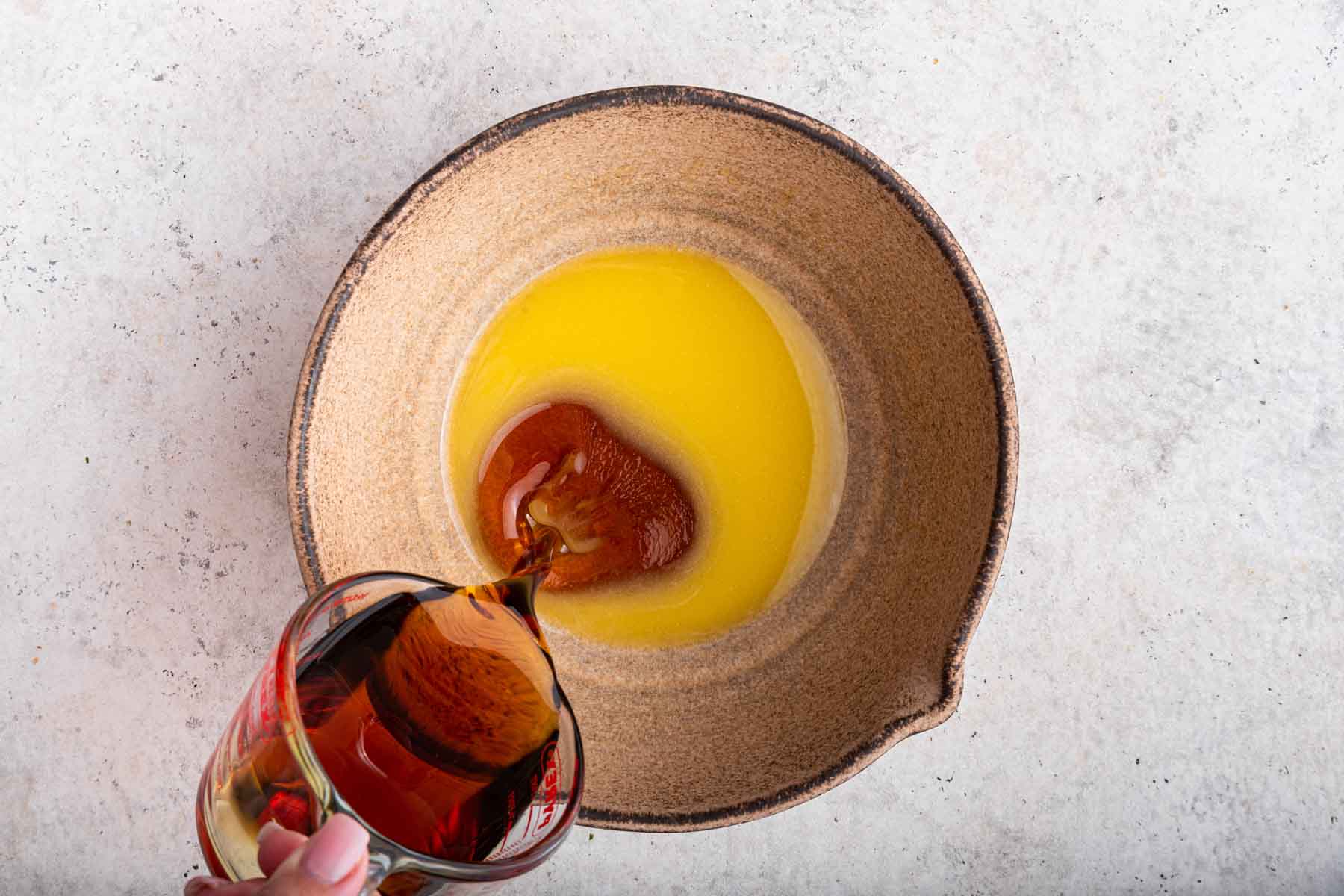 Pouring maple syrup into a yellow cake batter in a brown bowl.