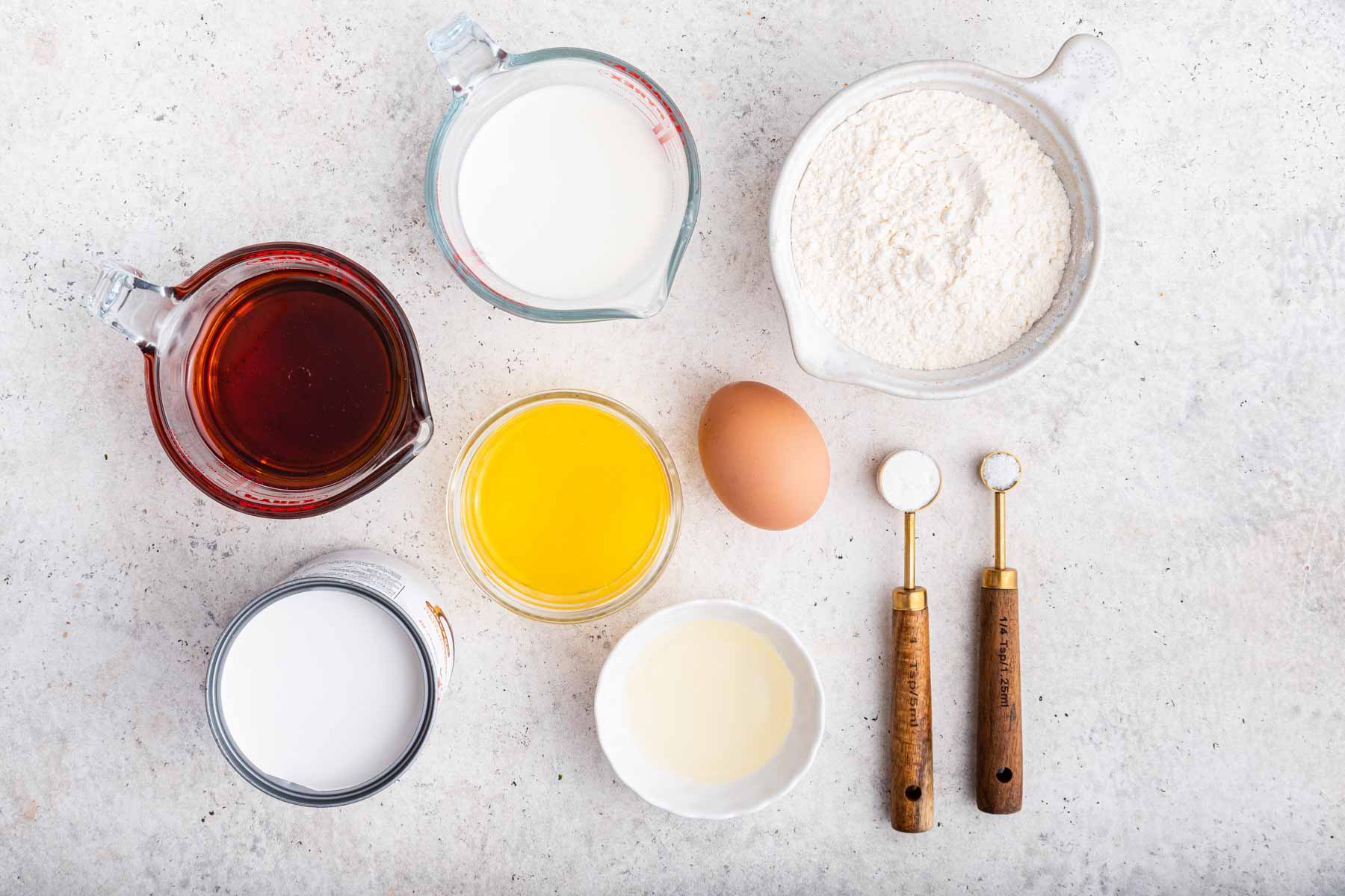 Bowls of maple syrup, flour, sugar, egg, and melted butter on grey counter.