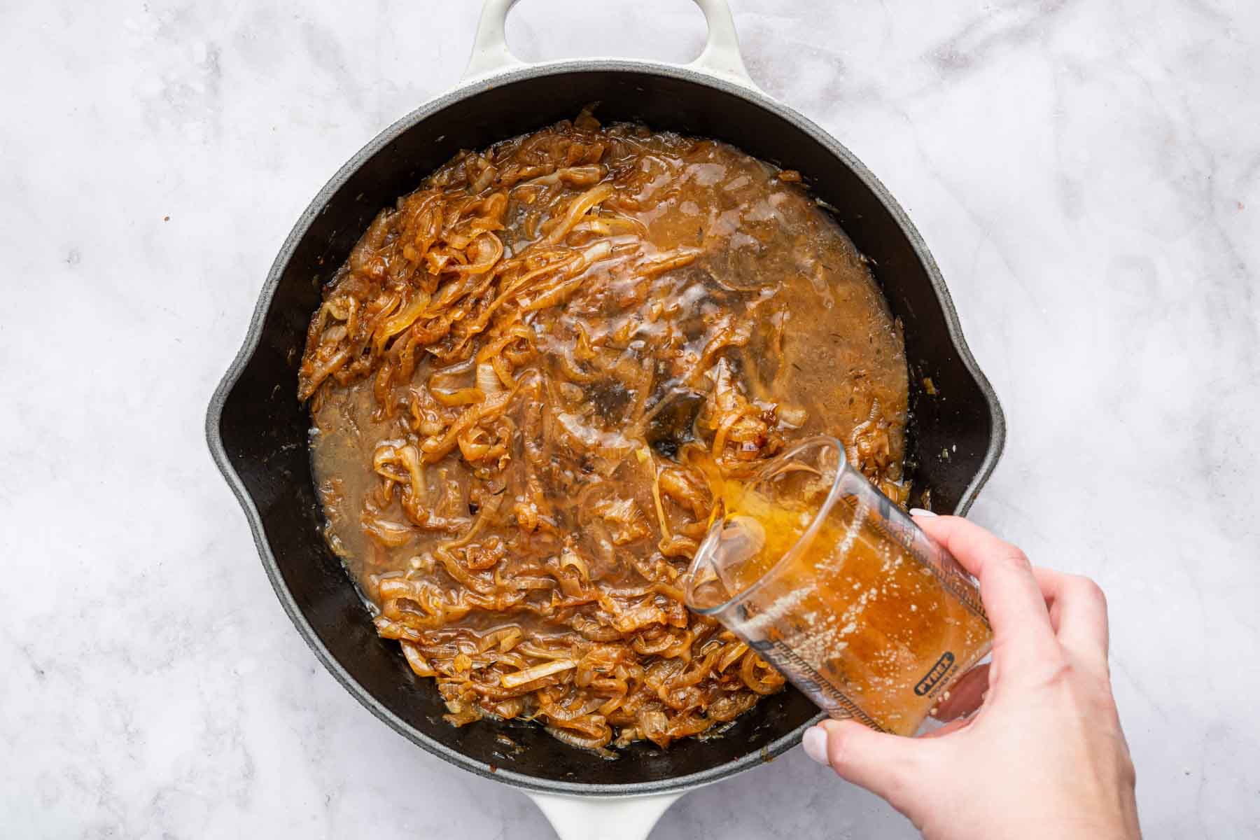 Hand pouring beef broth into cast iron skillet with cooked vegetables.