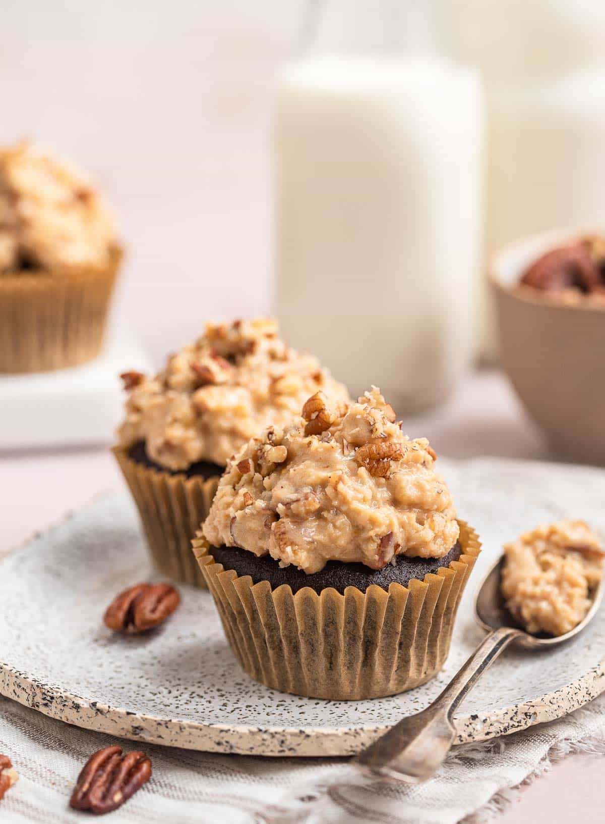 Two chocolate cupcakes on plate with spoon of frosting alongside.