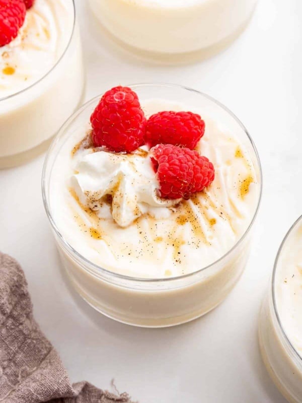 Vanilla pudding in small glass jars with raspberries and vanilla bean on top.