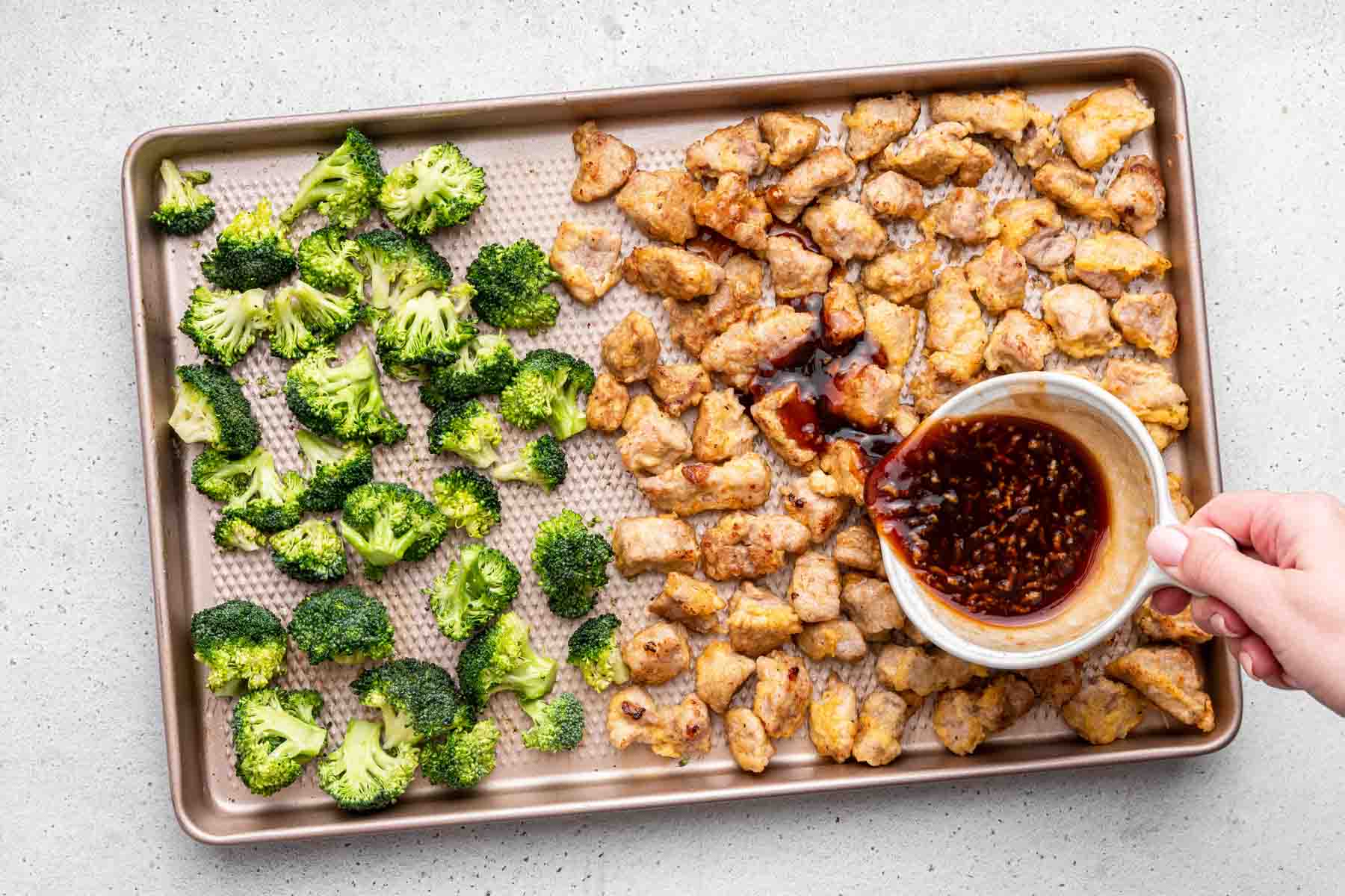 Sheet pan with broccoli on one side and brown pieces of fried meat on the other side with hand pouring sauce.