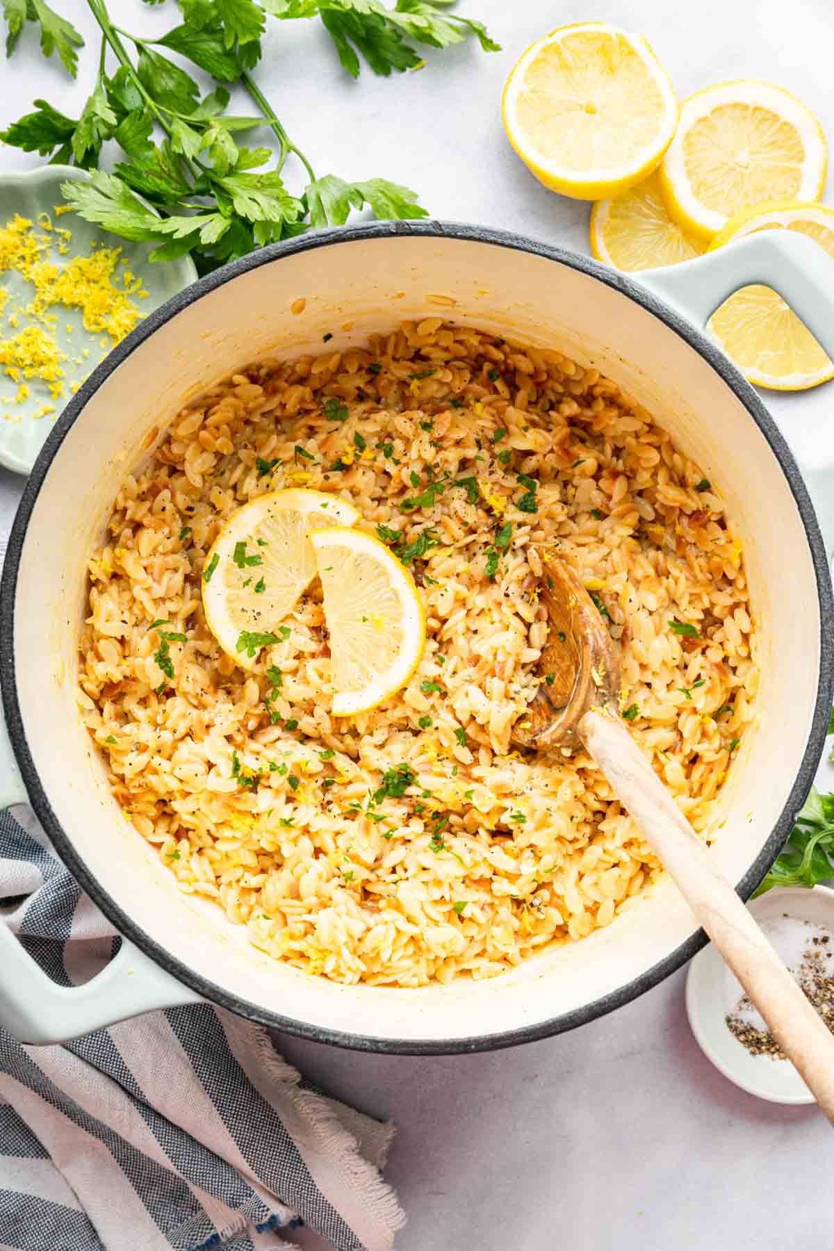 Lemon orzo in a white pan with a wooden spoon and lemon slices on the side.