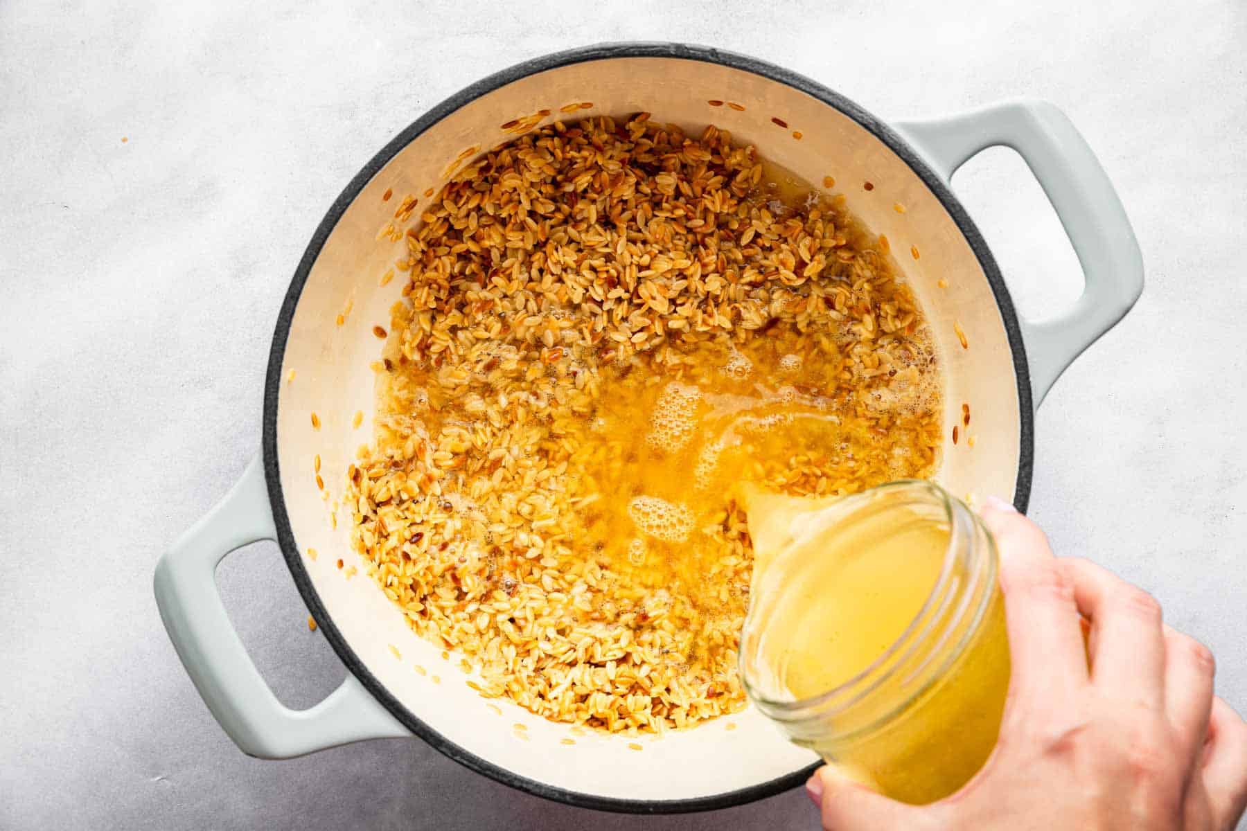 Hand pouring chicken broth into white pot with pasta.