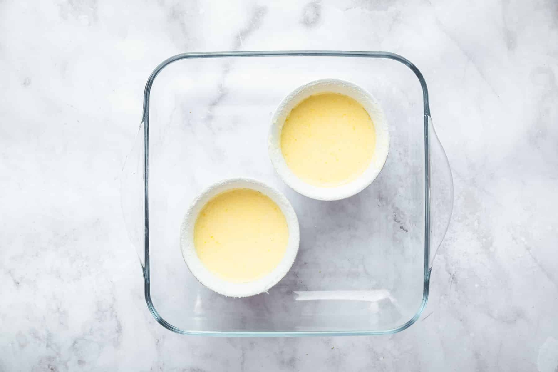 Two ramekins with pale yellow batter inside a square glass baking dish.