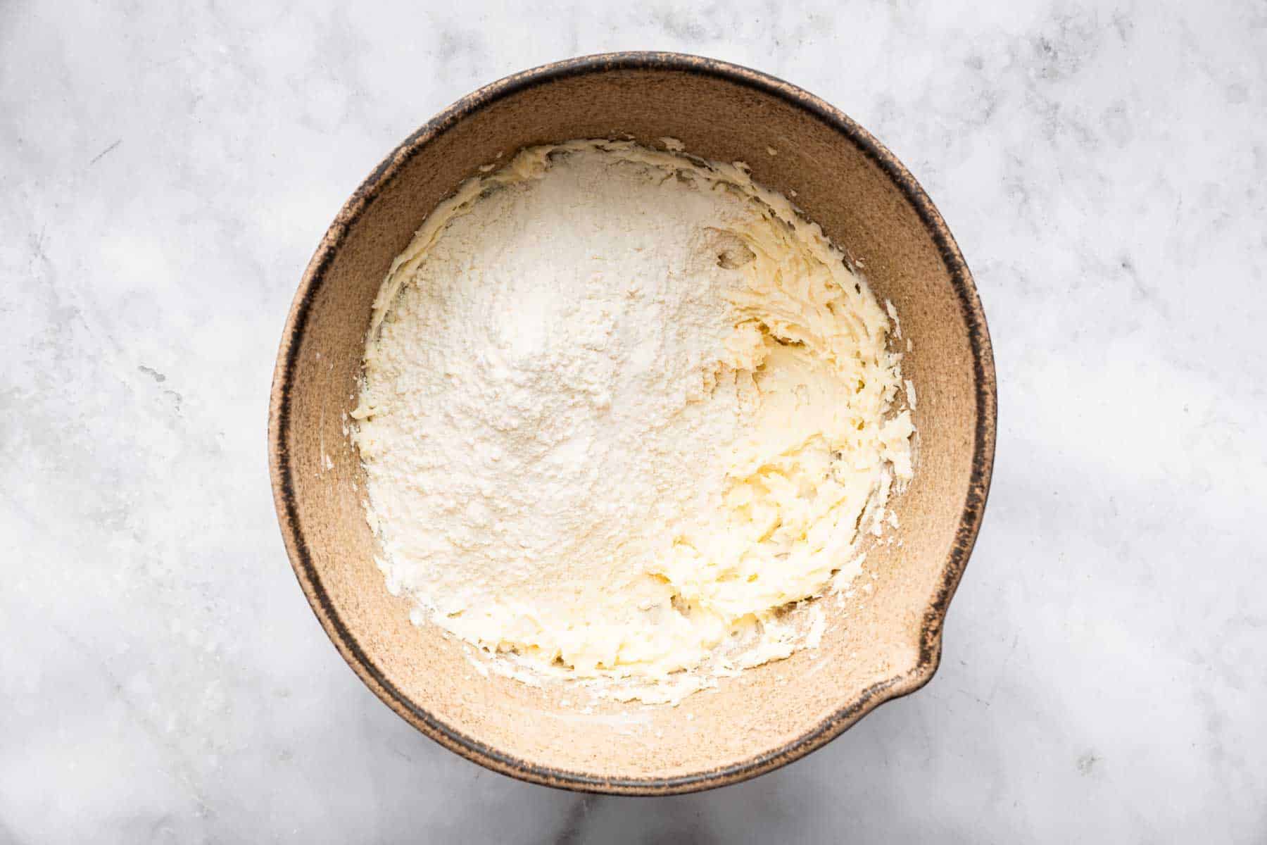 White flour sprinkled over bowl of pale yellow cookie dough in brown bowl on marble counter.