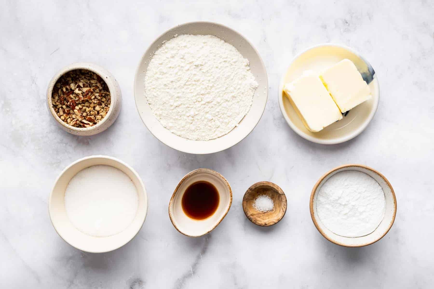 White bowls filled with flour, diced butter, vanilla, and chopped pecans.