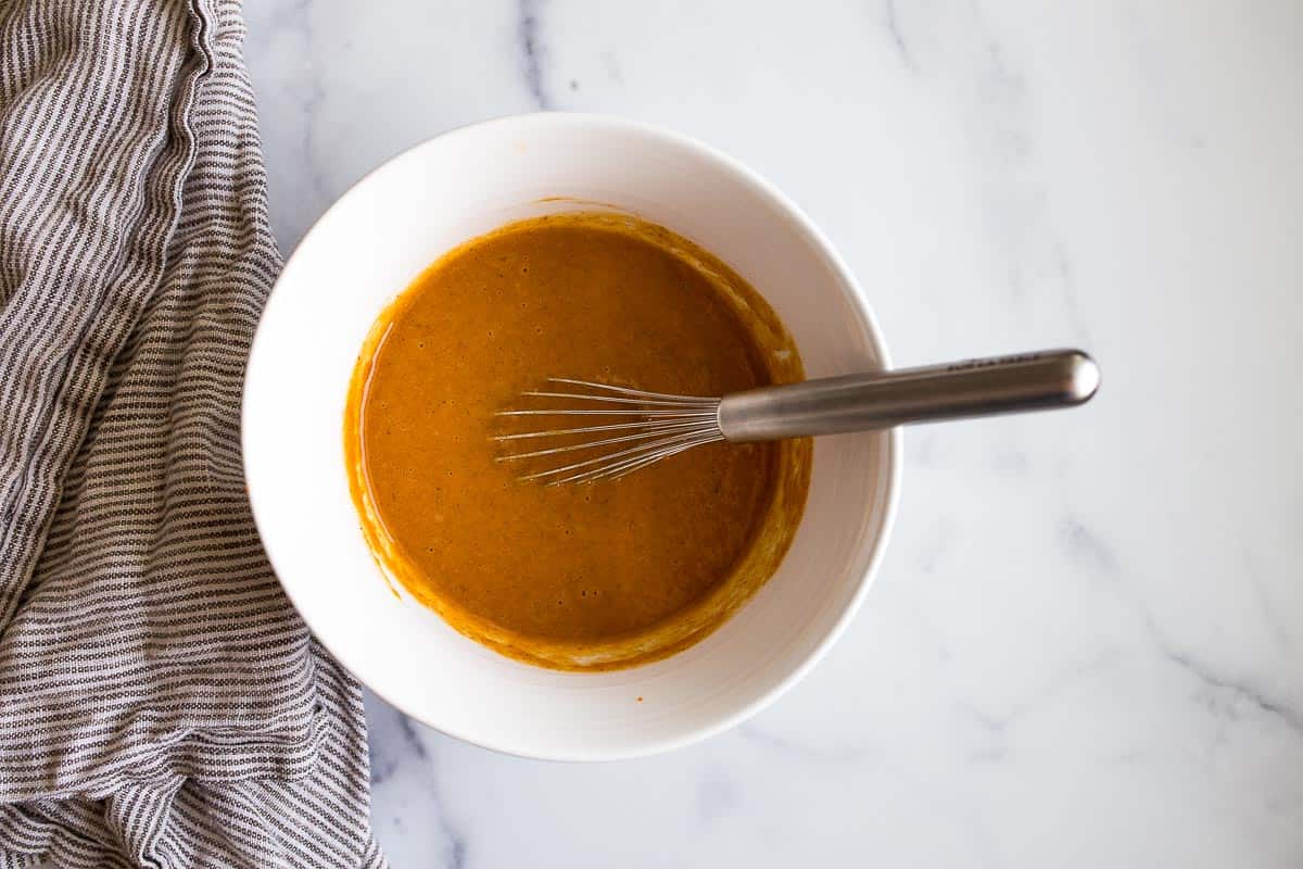 White bowl of dark orange liquid batter with whisk in it.
