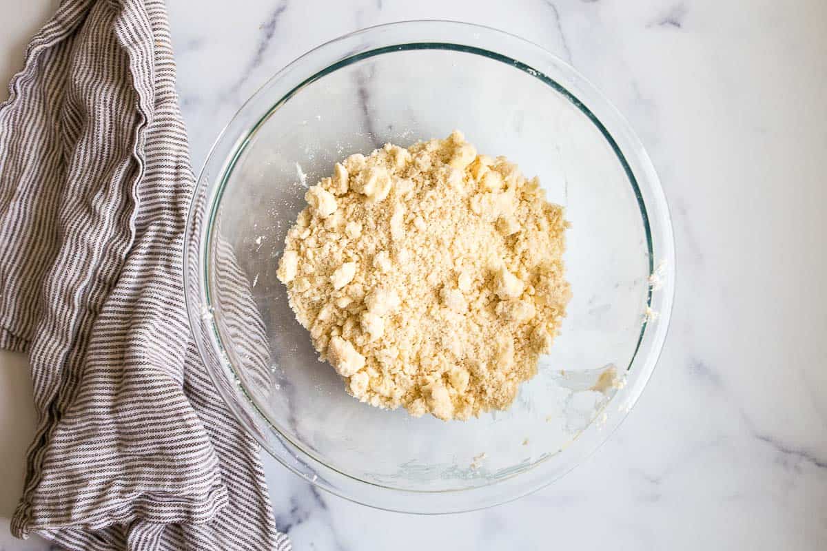 Crumbly yellow dough in a clear glass bowl on marble counter.
