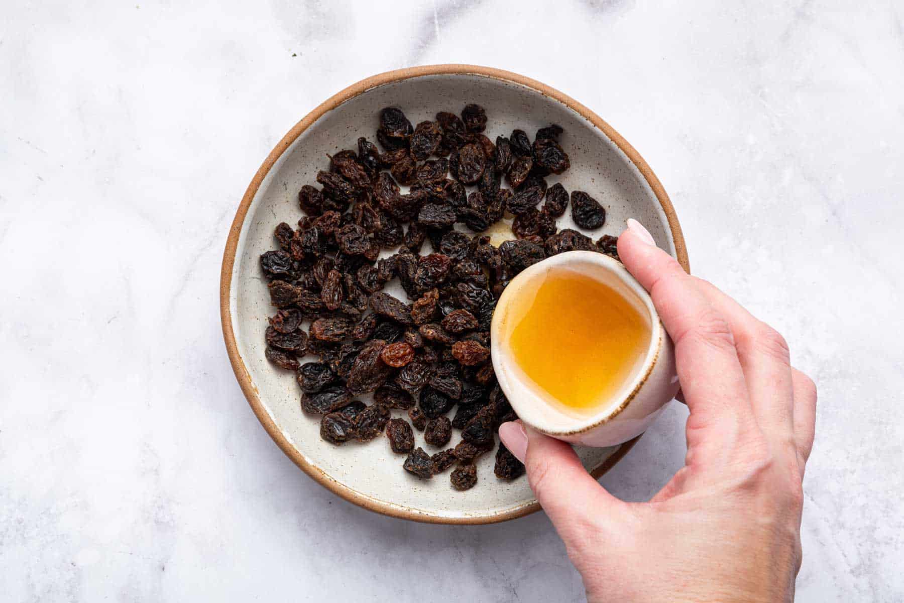 Raisins in a bowl with a hand pouring amber liquid on top.