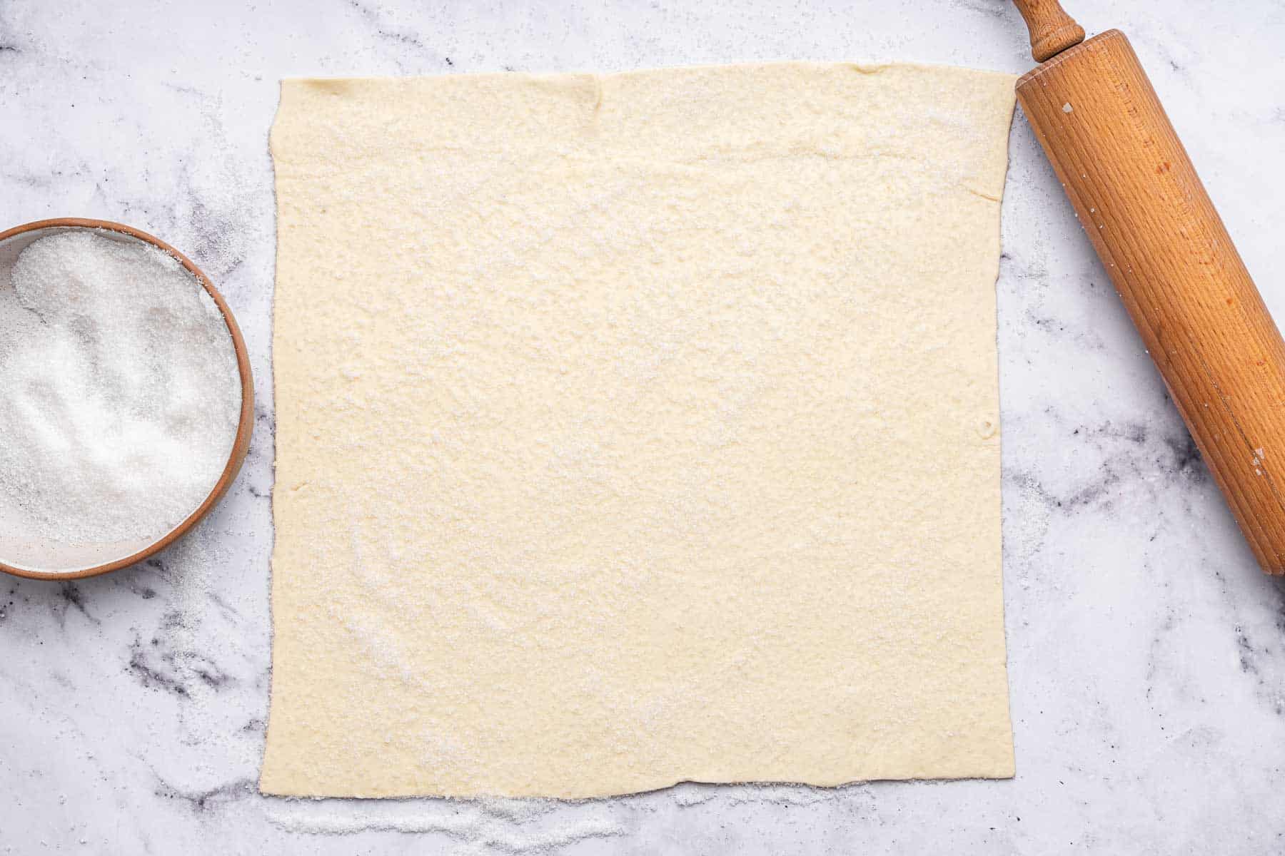 Rectangular piece of pastry on marble surface with rolling pin on side.