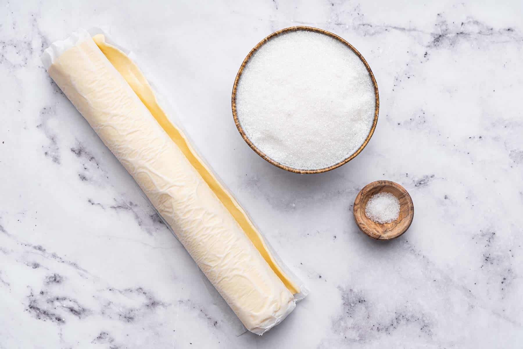 Horizontal photo of a roll of puff pastry, a bowl of sugar and salt on marble surface.