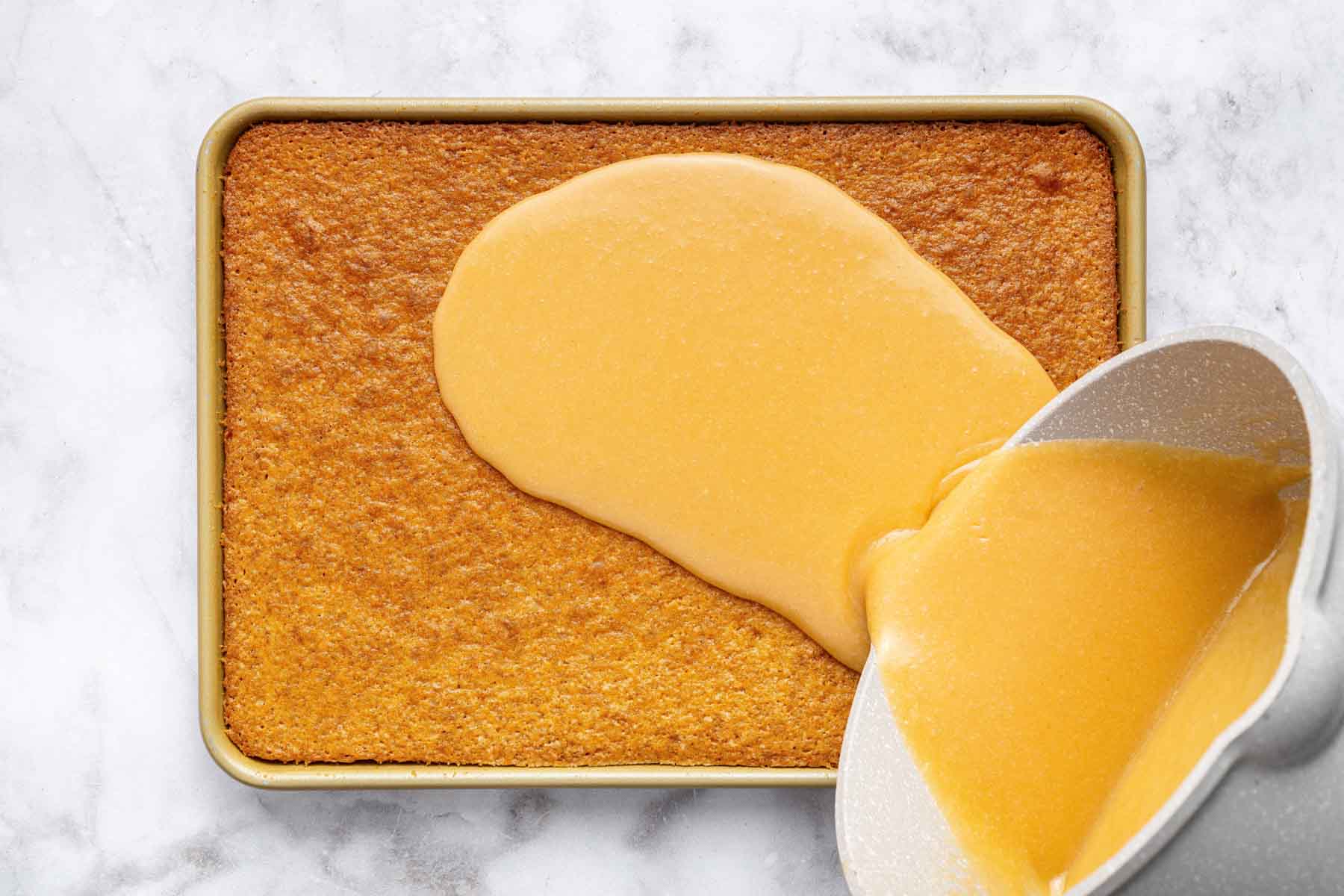 Runny brown frosting being poured on golden brown rectangular cake.