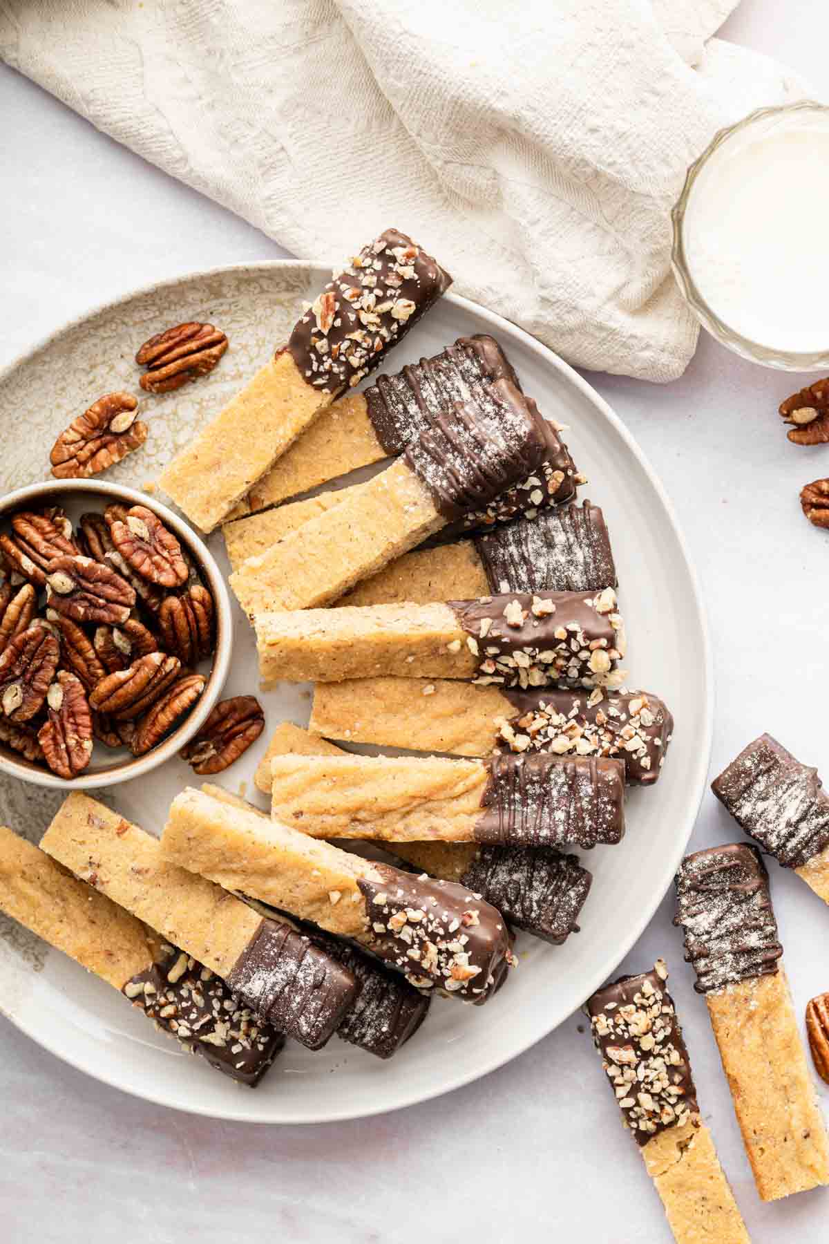 Vertical image of pecan shortbread cookie sticks on a plate with a bowl of pecans.