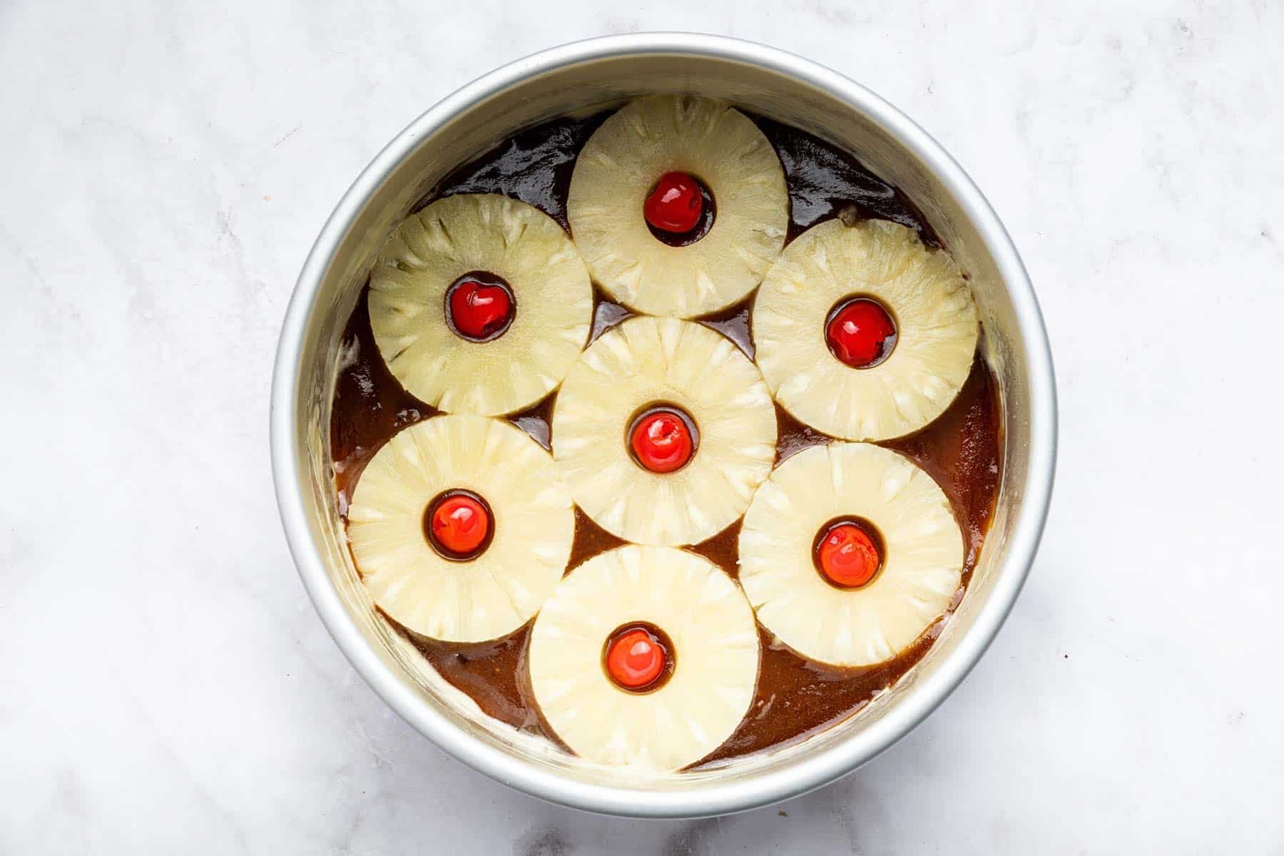 Seven pineapple rings with cherries in the center over brown caramel in a round cake pan.