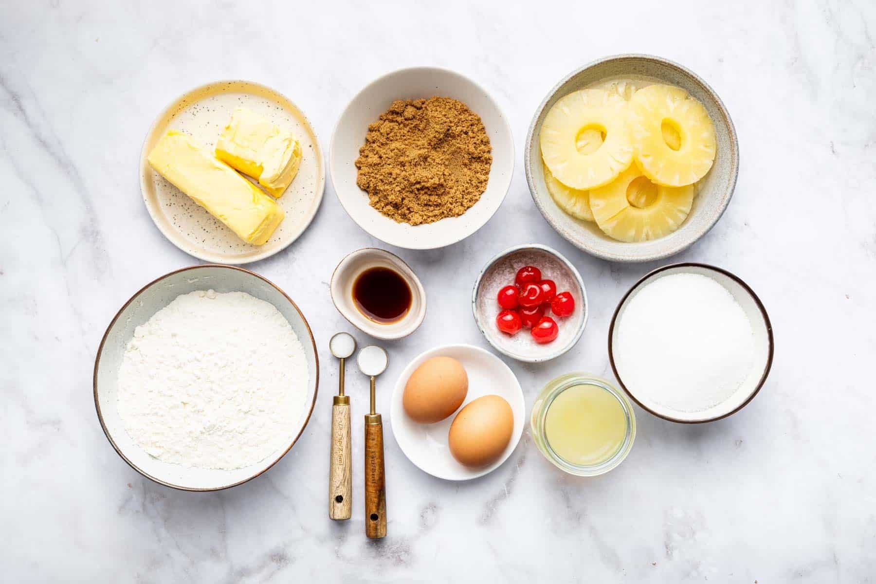 Bowl with fruit rings, cherries, eggs, flour, butter and vanilla on kitchen counter.