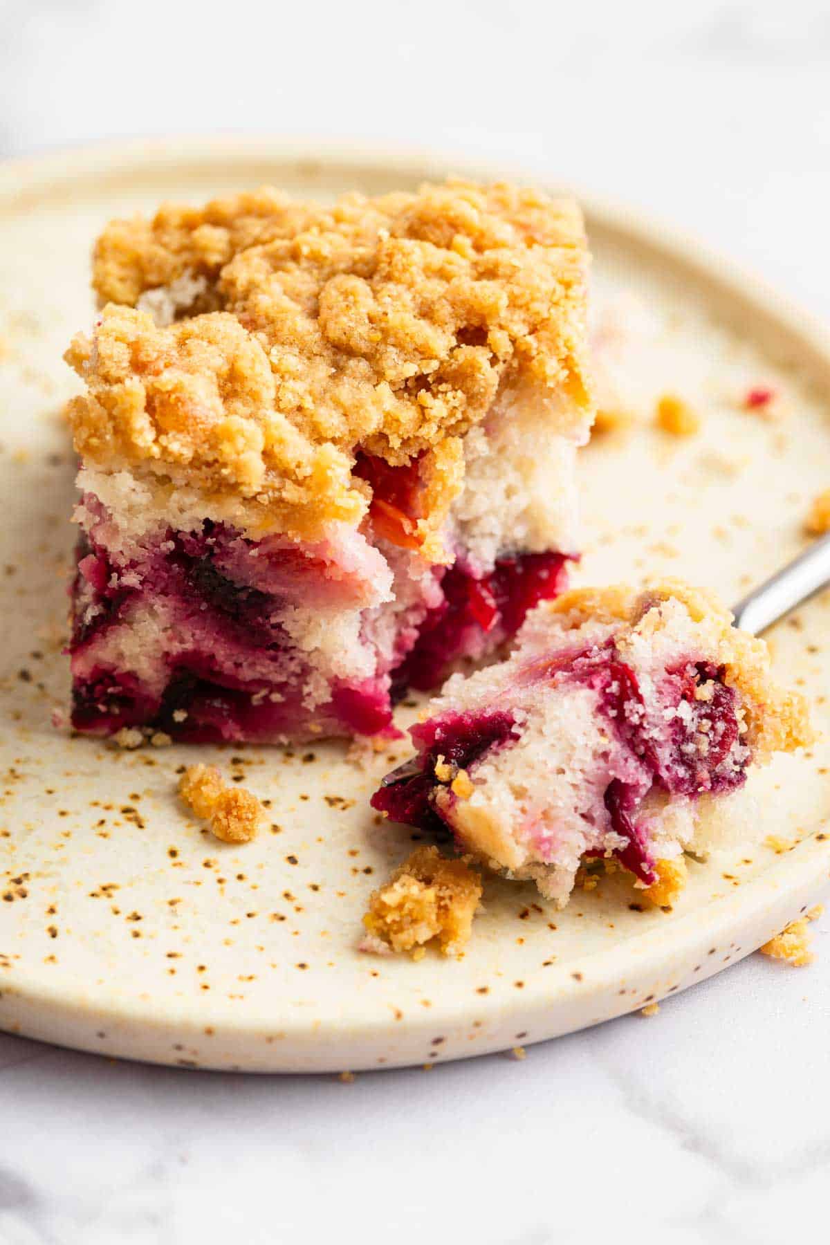 Square slice of plum cake on plate with fork removing a bite.