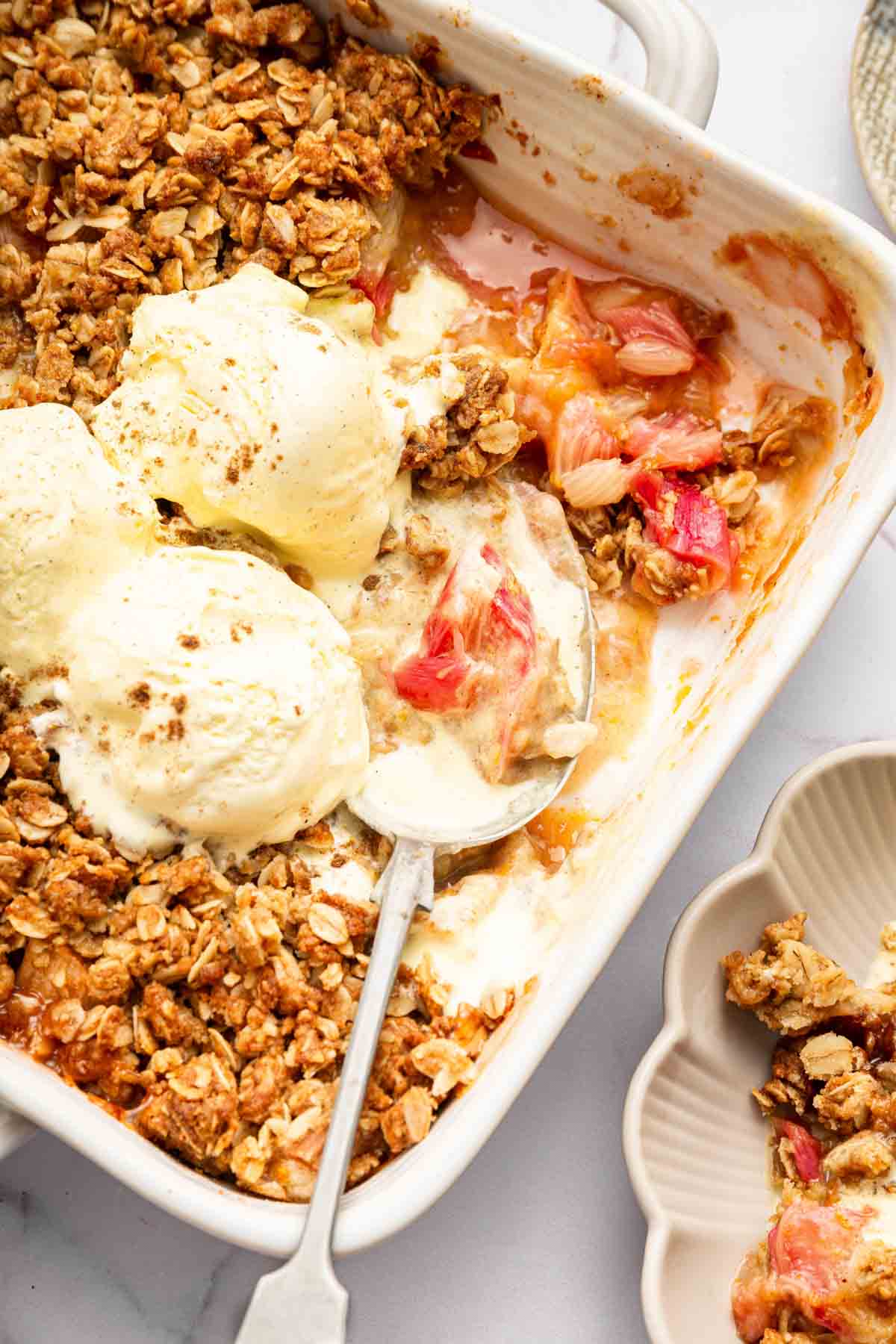 Vertical image of rhubarb crisp with spoon in it and ice cream melting on top.
