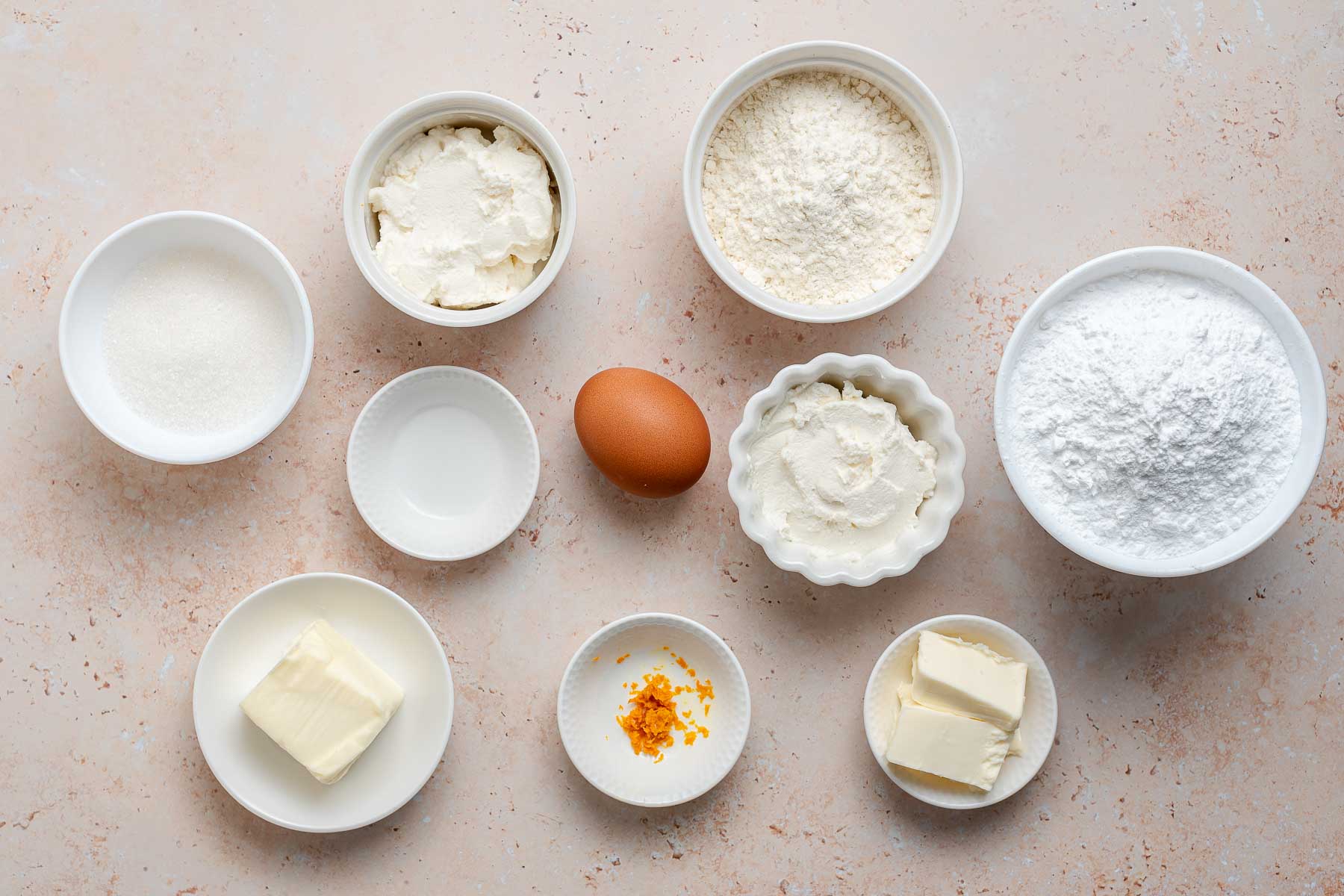 Ingredients to make cupcakes in little bowls on a light pink table.