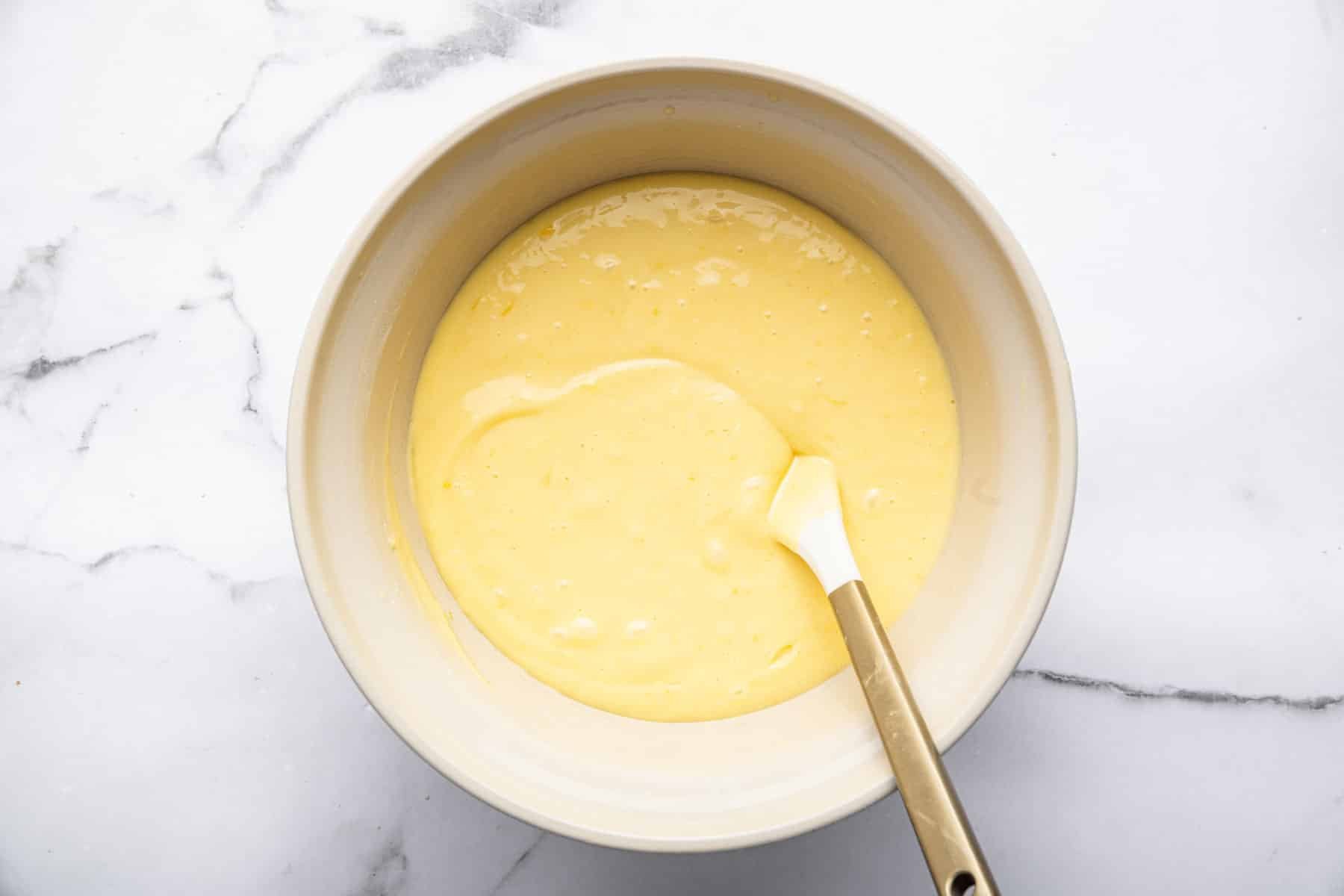 Cream colored bowl with yellow batter and white spatula.