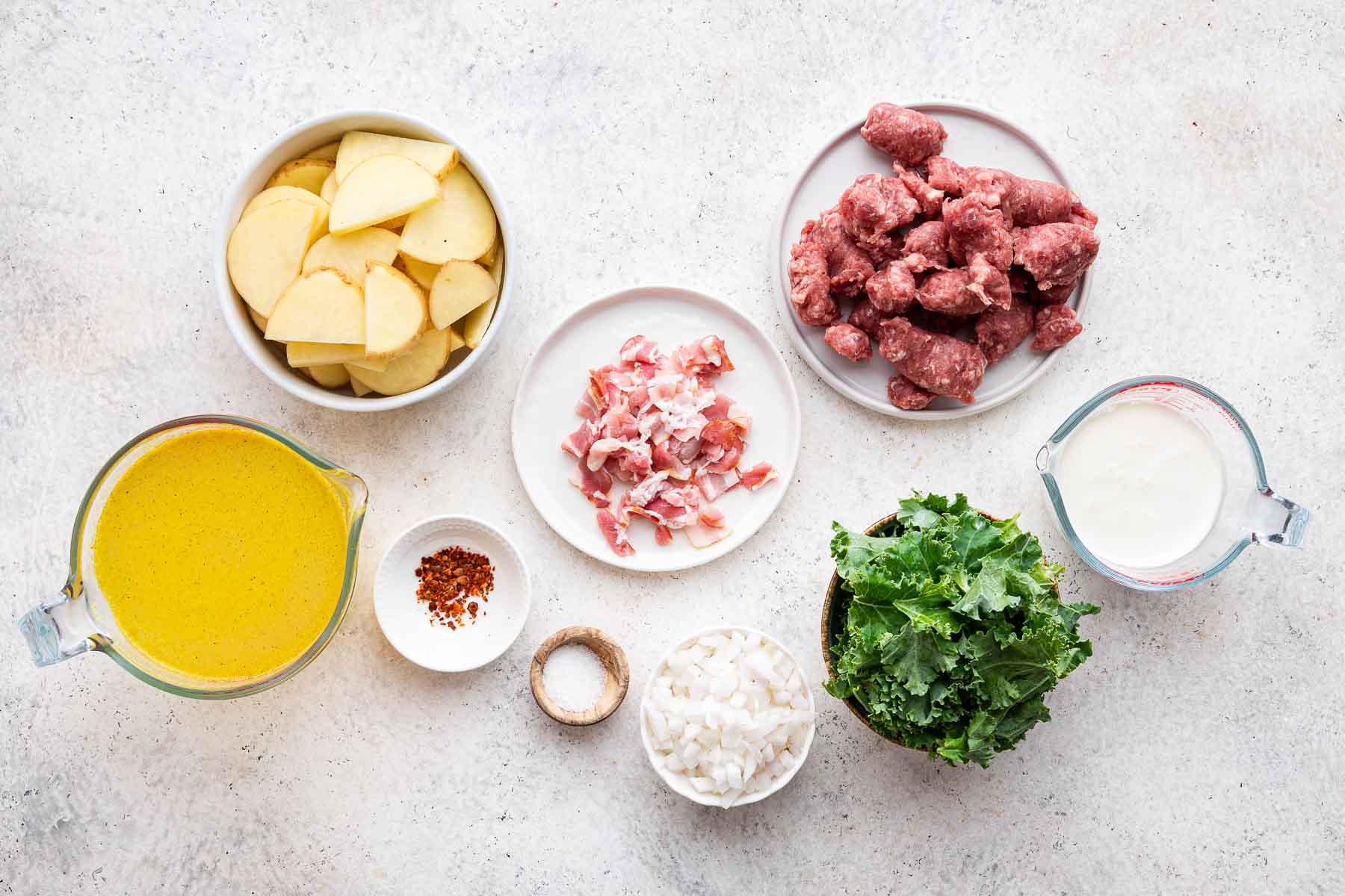 Small bowls of sausage, kale, chicken broth, bacon, and sliced potatoes on white counter.